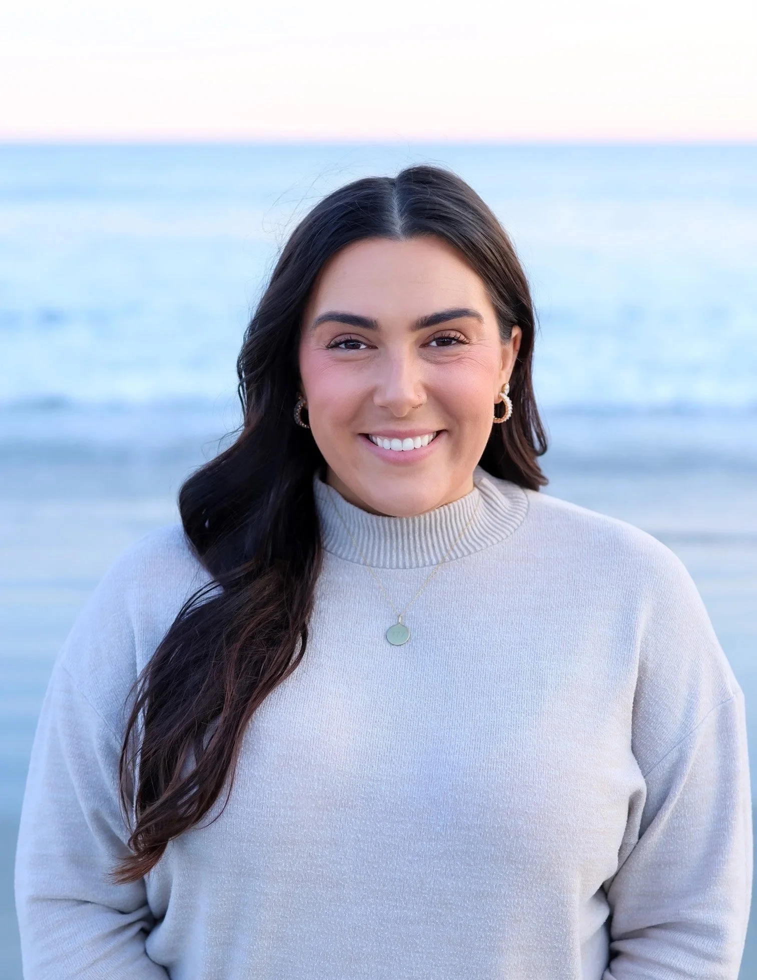 A woman smiling at the camera standing by the beach with waves in the background.