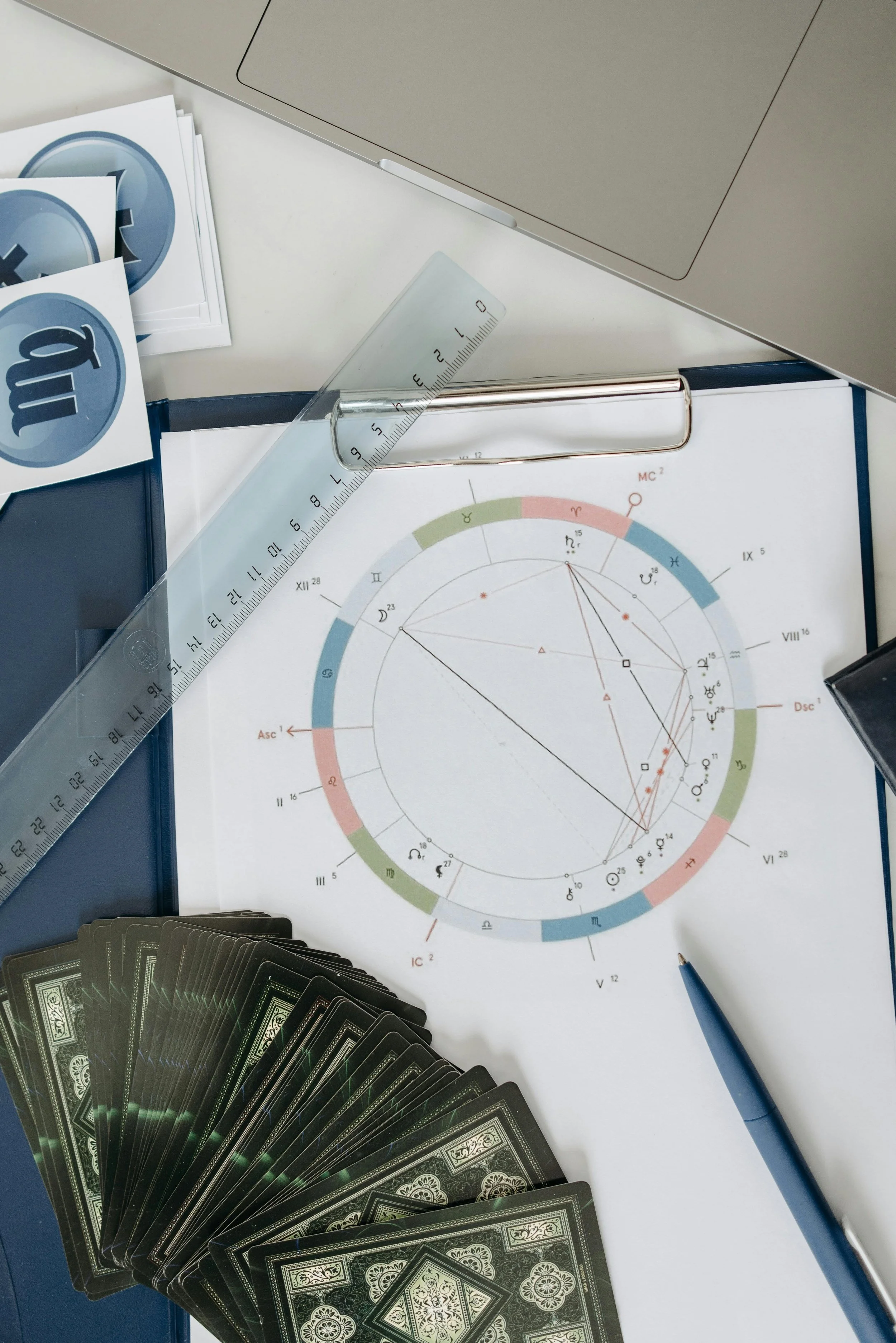 An astrology chart with various symbols and lines, a deck of playing cards, a clear ruler, a pen, and some stickers on a desk.