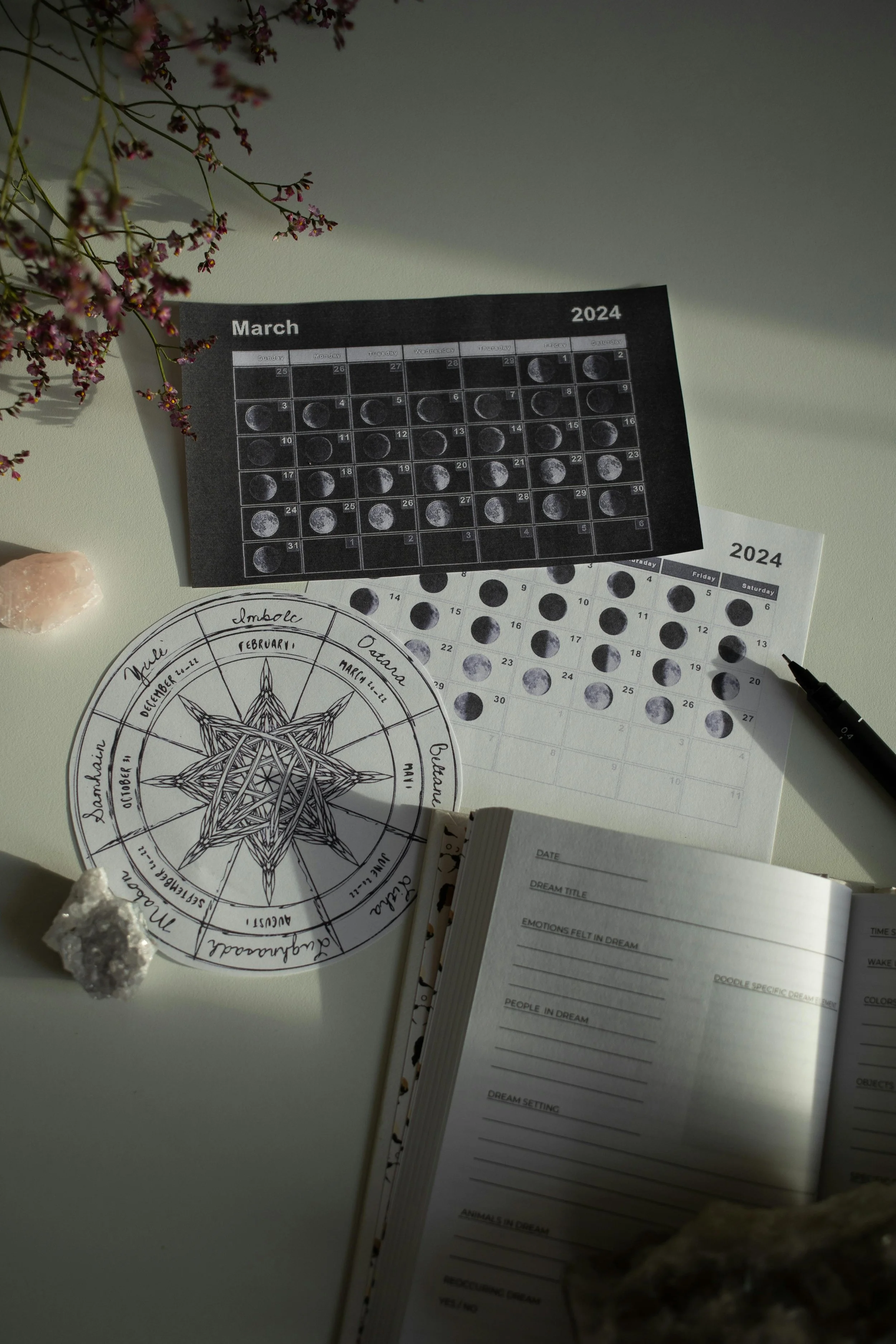 A desk setup with a lunar calendar for March 2024, a moon phase chart, an open dream journal, and lunar phase stickers, surrounded by rocks and flowers.