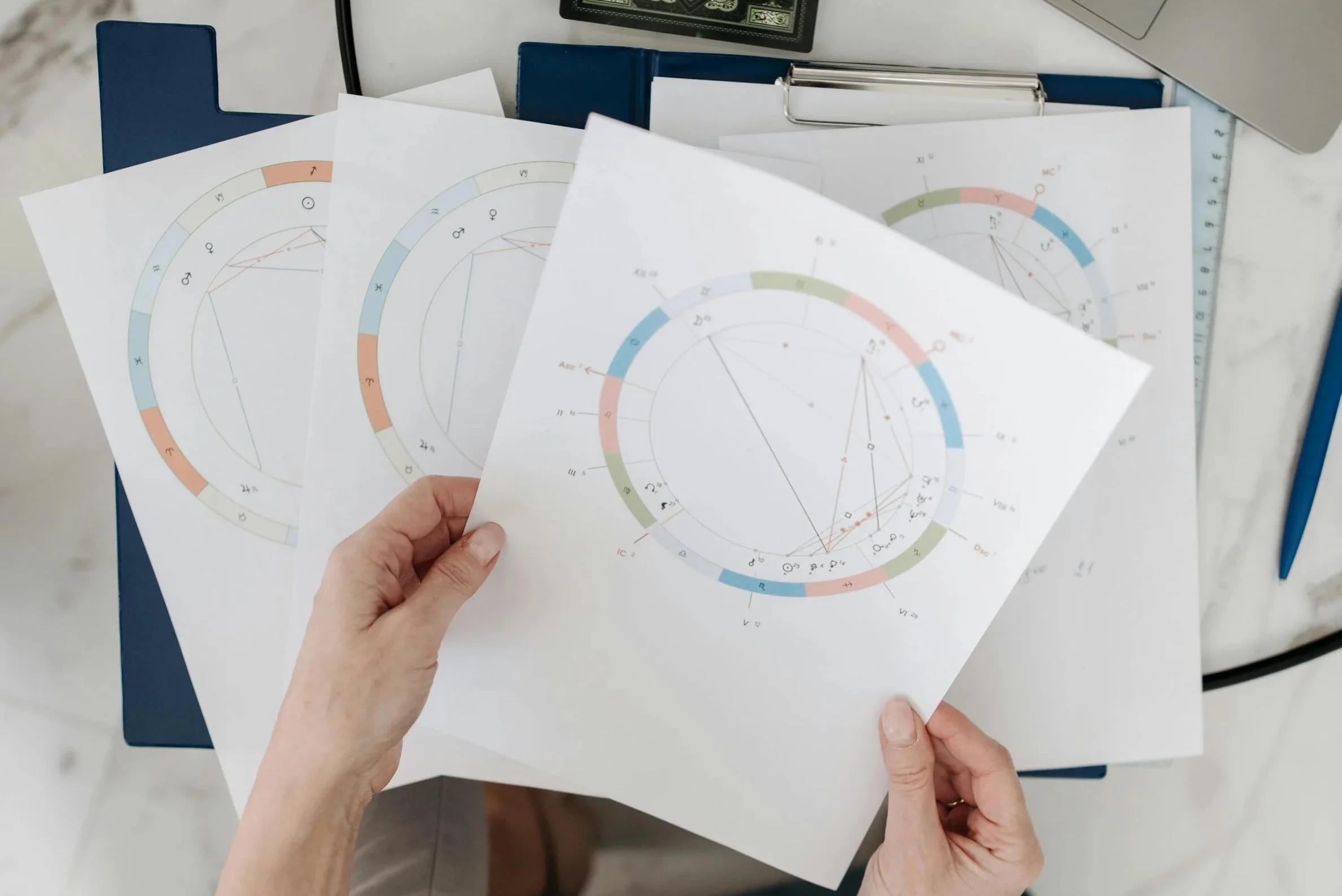 Person holding printed astrological charts with zodiac symbols and geometric lines, placed on a desk with a blue folder, a clipboard, and a computer.
