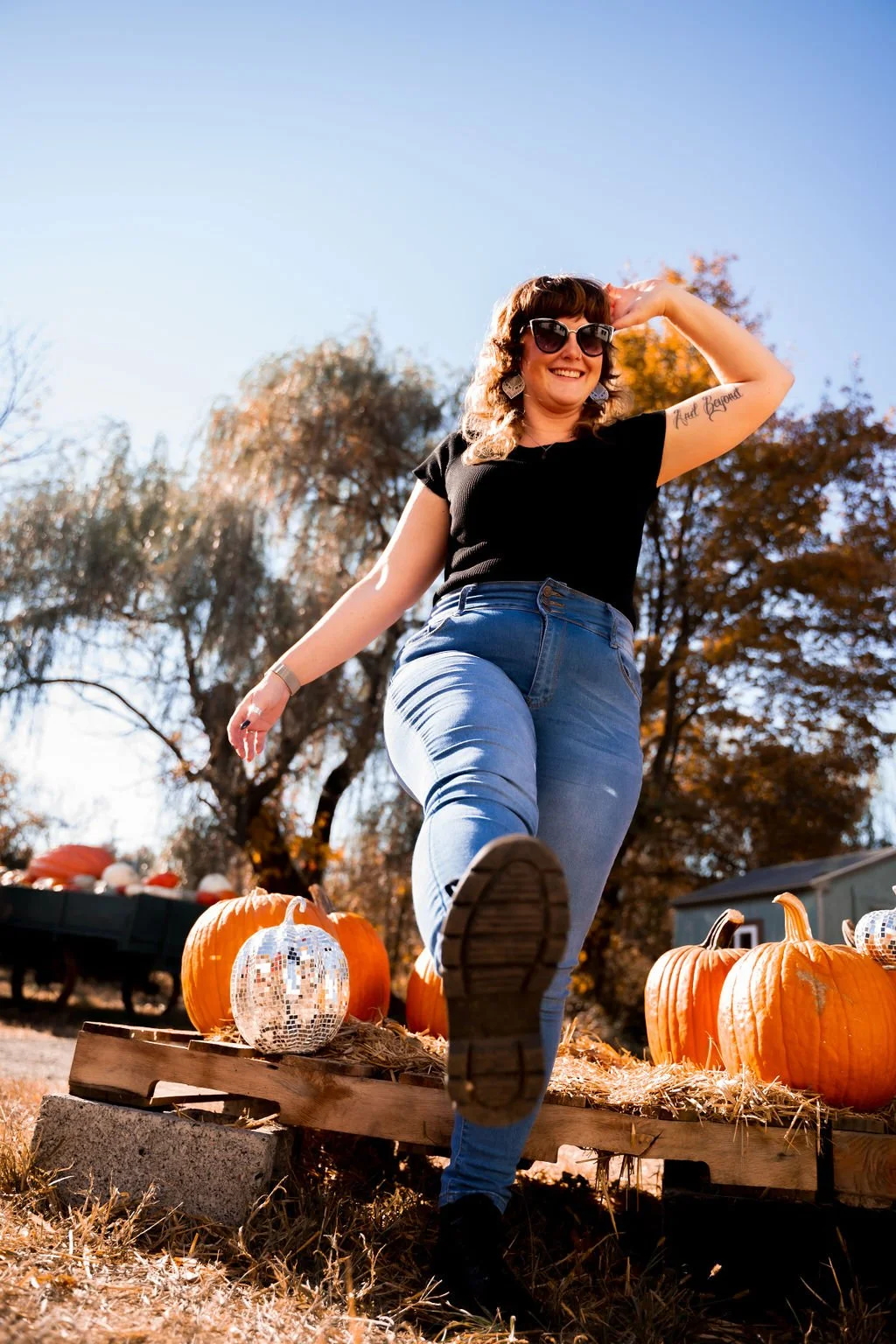 Candid photo of Felicia, sunglasses on foot and arm up in front of pallet of pumpkins