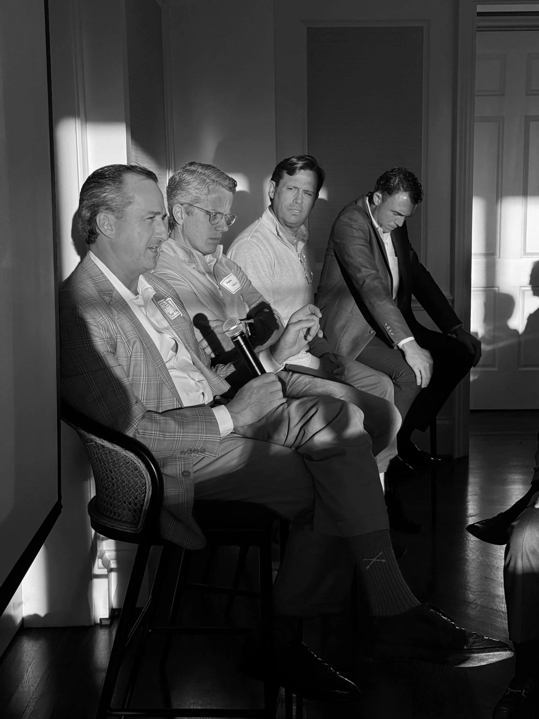 Black and white photo of four men sitting against a wall; one holding a microphone, appearing to be in a panel or interview setting, with sunlight casting shadows on the wall.