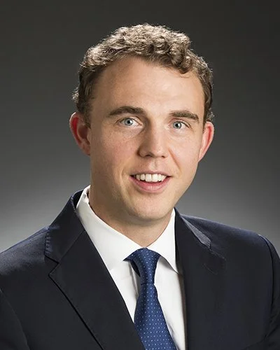 Professional portrait of a young man with curly blonde hair, wearing a dark suit, white shirt, and blue tie, smiling against a gray background.