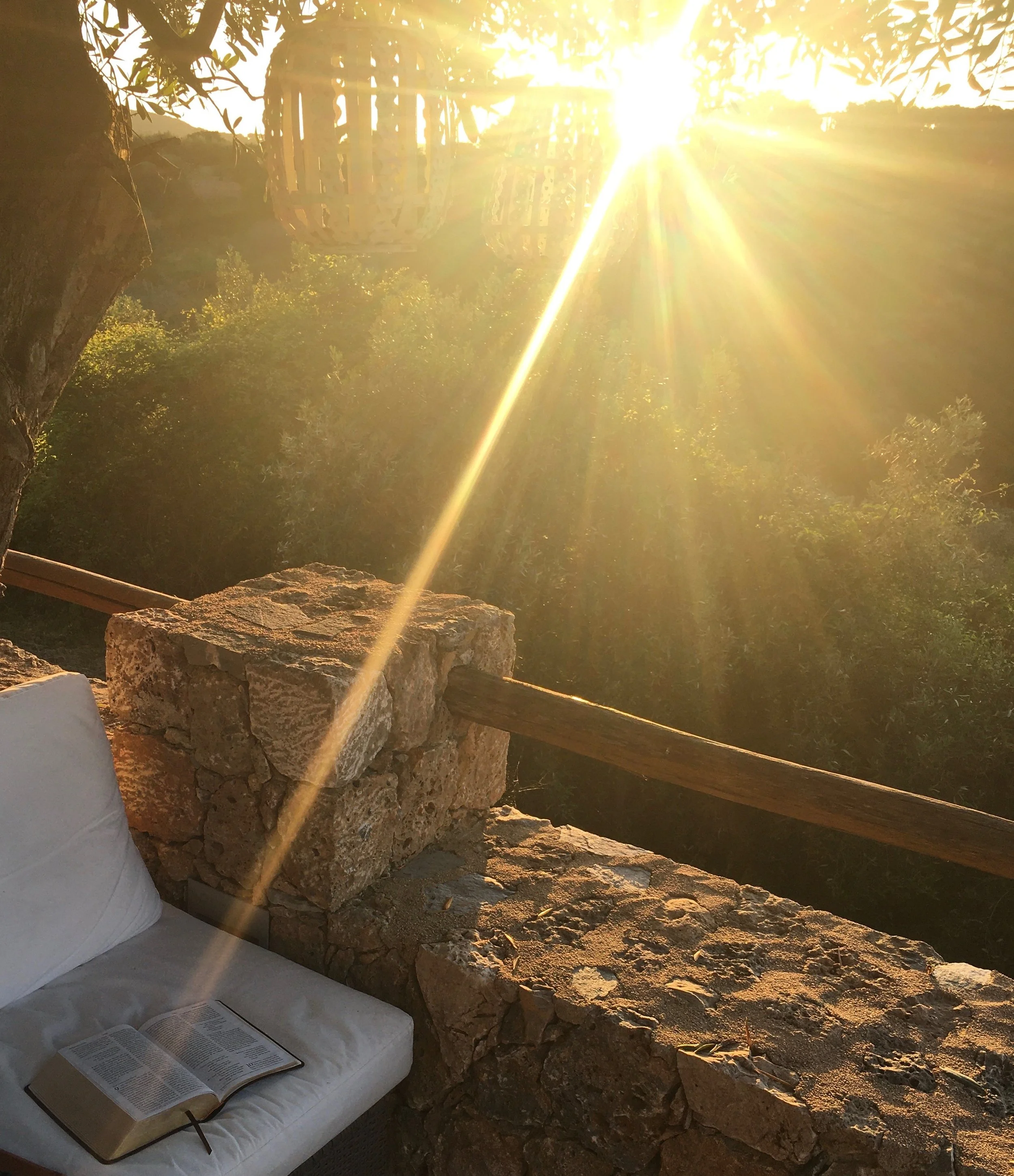 Sunlight shining through trees onto a stone and wood balcony with a white cushion and an open book.