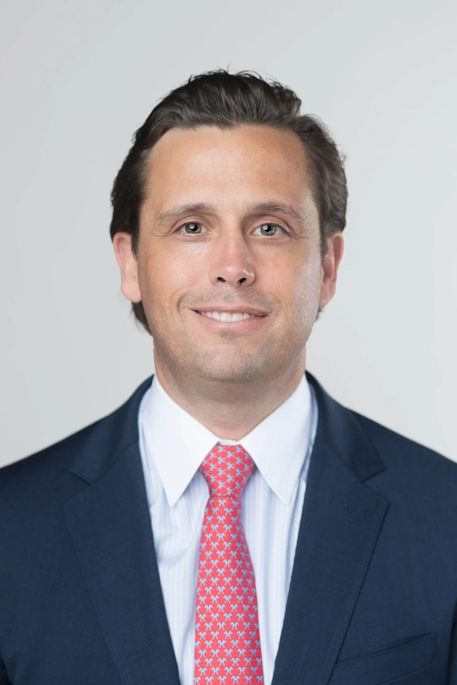 A professional portrait of a man in a dark blue suit, white shirt, and red patterned tie, smiling against a neutral background.