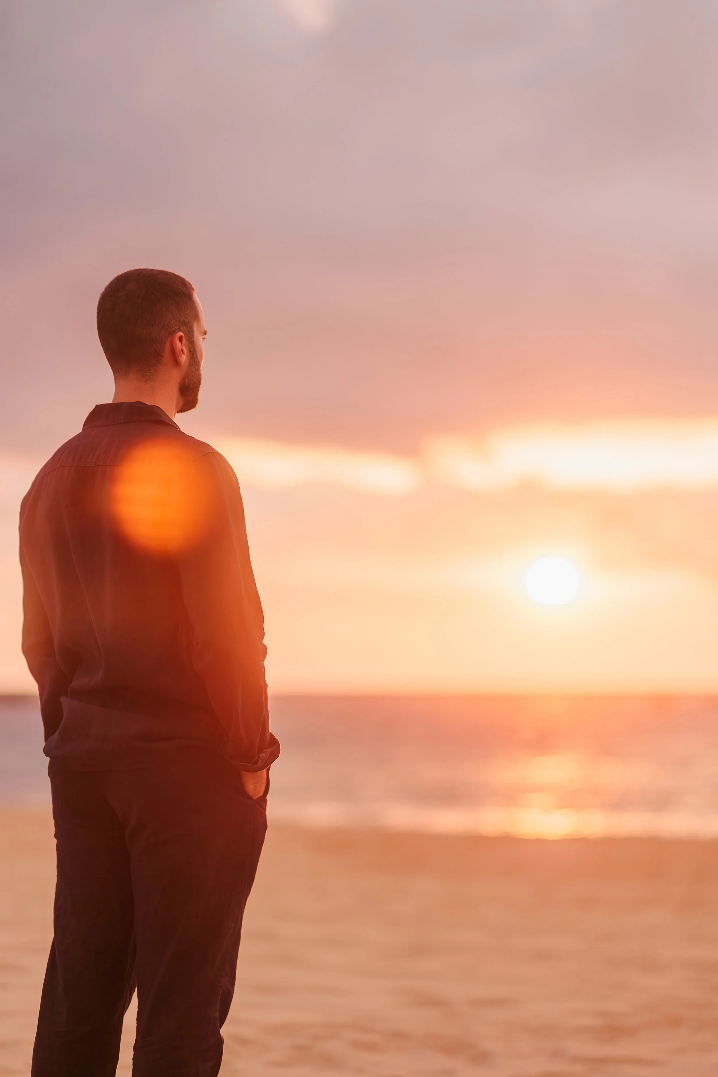 A man standing by the beach during sunset, looking at the horizon with his hands in his pockets.