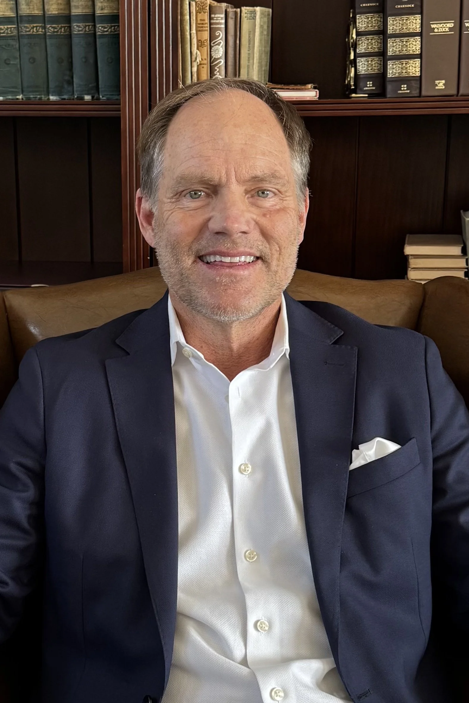 A middle-aged man with short brown hair and facial stubble, wearing a dark blue suit jacket and a white dress shirt, sitting on a brown leather chair in front of a wooden bookshelf filled with books.