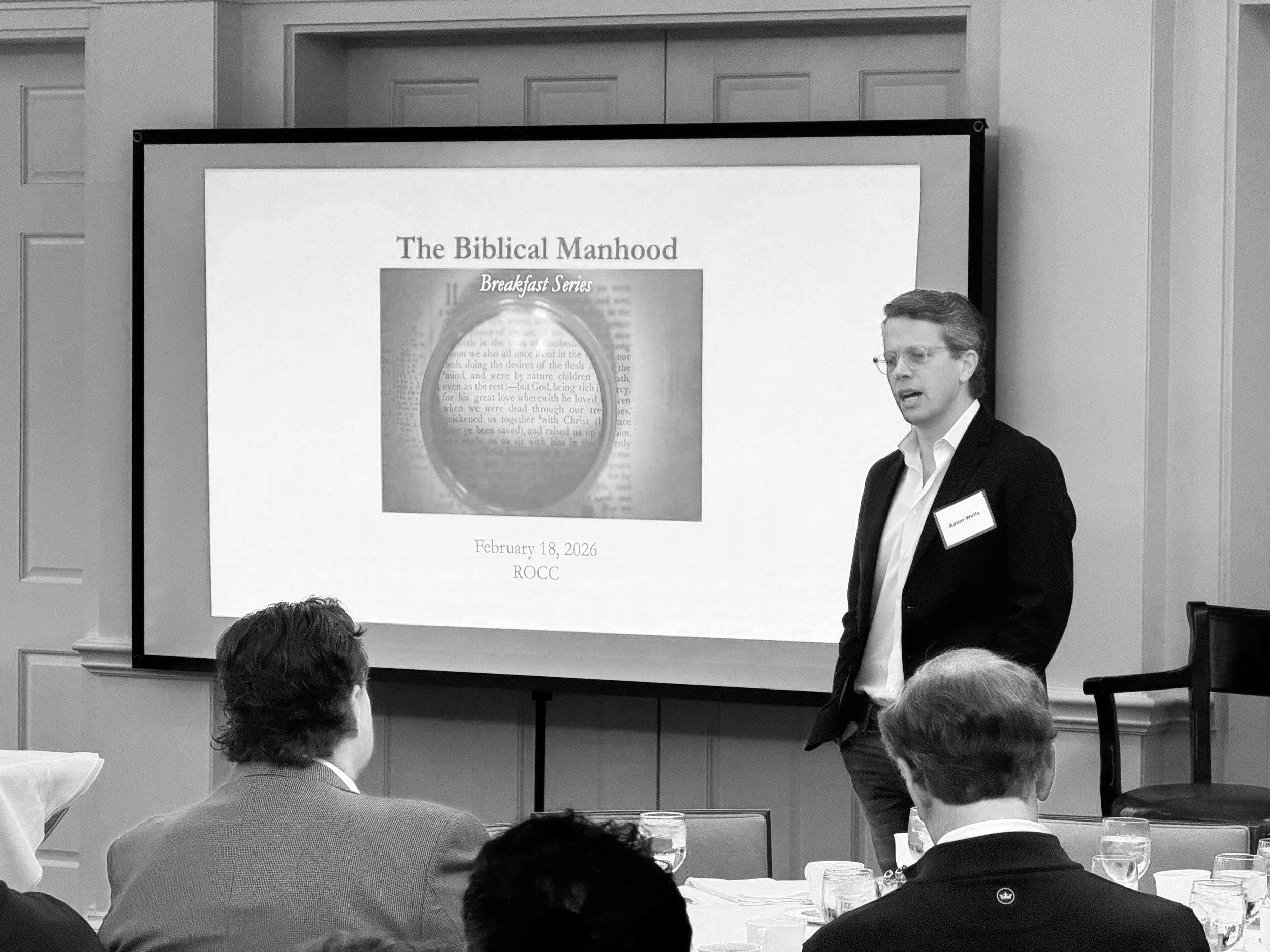 A man giving a presentation on biblical manhood at a conference, standing beside a large screen displaying a slide titled 'The Biblical Manhood'. The slide shows a cover of a book called 'Breakfast Series' with the subtitle and date February 18, 2026, ROCC. The audience is seated at tables with drinks.