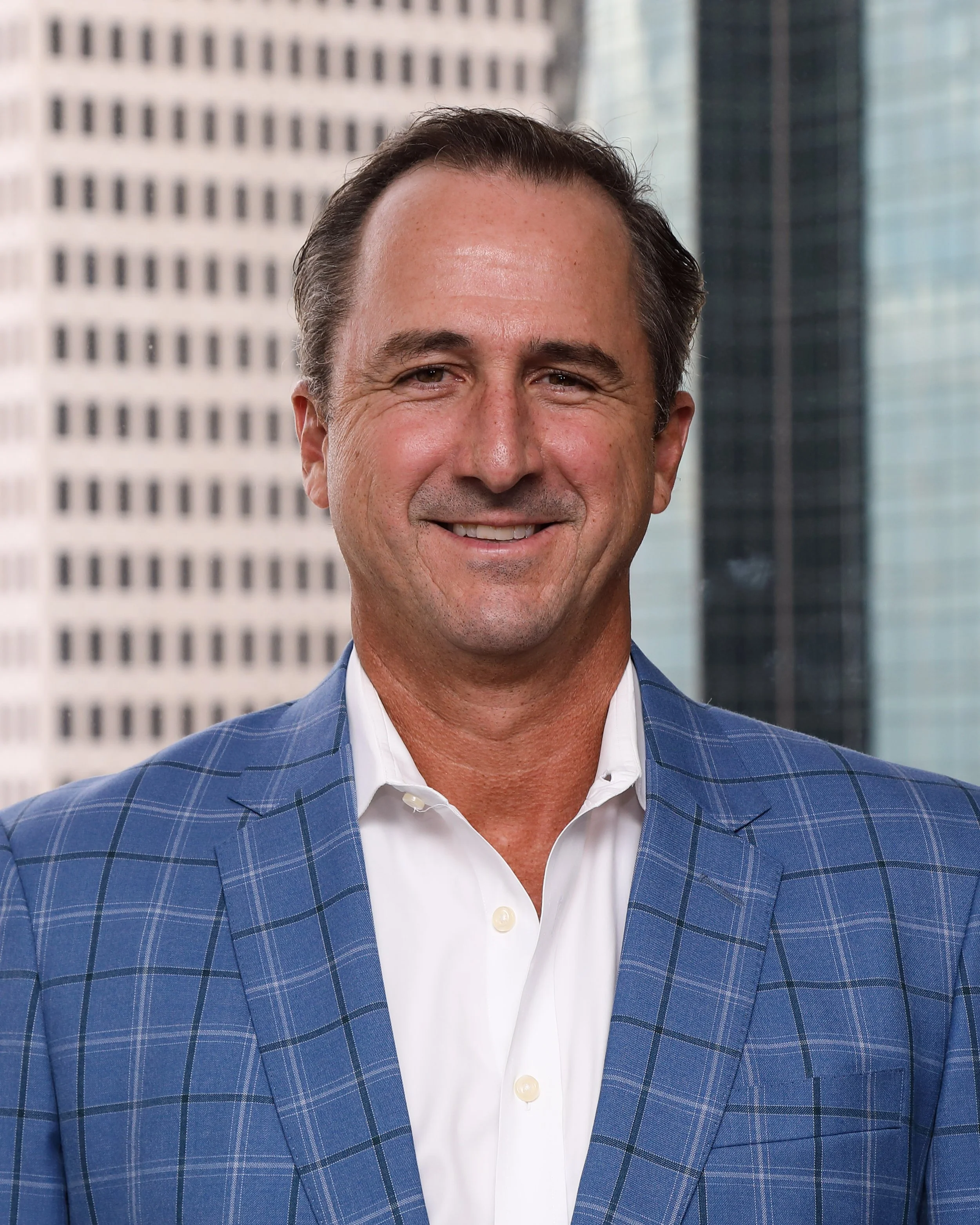 A smiling man with dark hair, wearing a blue plaid blazer and a white collared shirt, stands in front of modern glass and concrete office buildings.