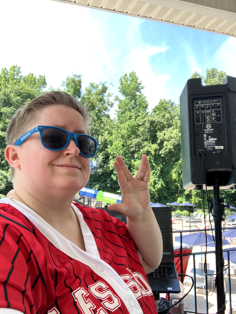 Ashley gives the "Live Long and Prosper" hand sign at an outdoor pool party. She is wearing a red and white baseball shirt and wearing blue sunglasses.