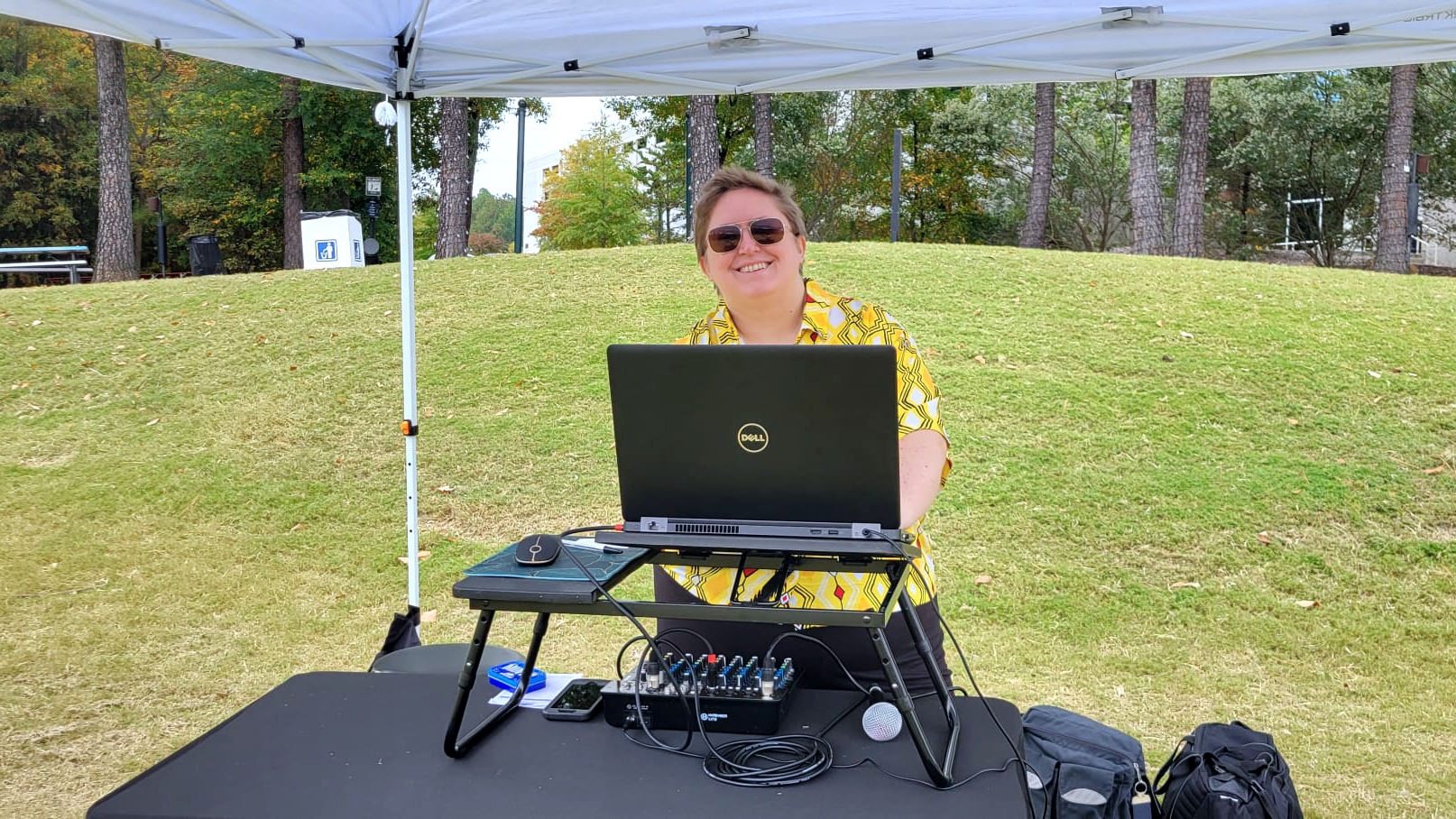 Ashley behind her console at a local outdoor event, wearing a yellow patterned shirt and sunglasses.
