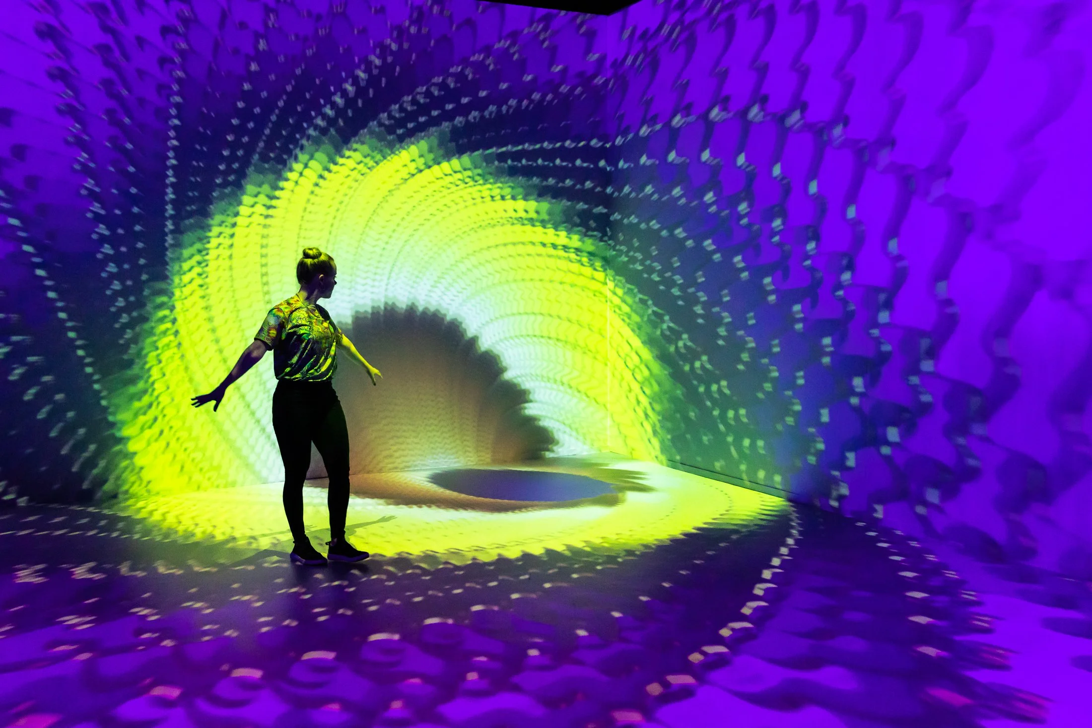 A person standing in a vibrant, abstract light installation with swirling purple and yellow patterns projected onto the walls and floor.