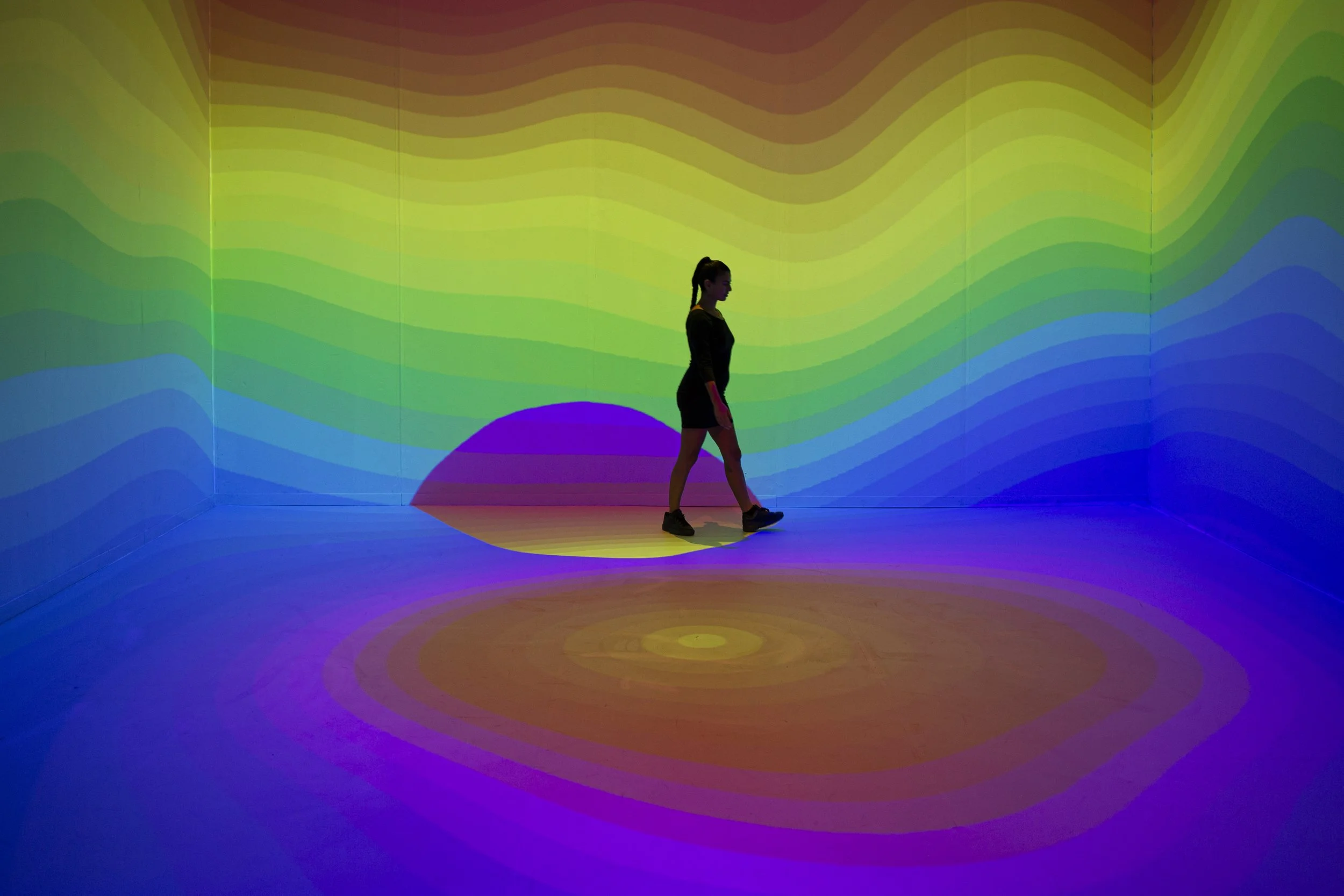 A woman walking across a room with colorful, wavy light projections on the walls and floor.