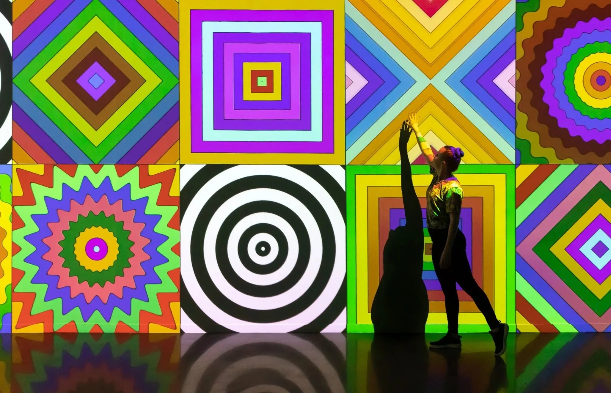 Silhouettes of two people dancing in front of a colorful, geometric, optical illusion-style light display.
