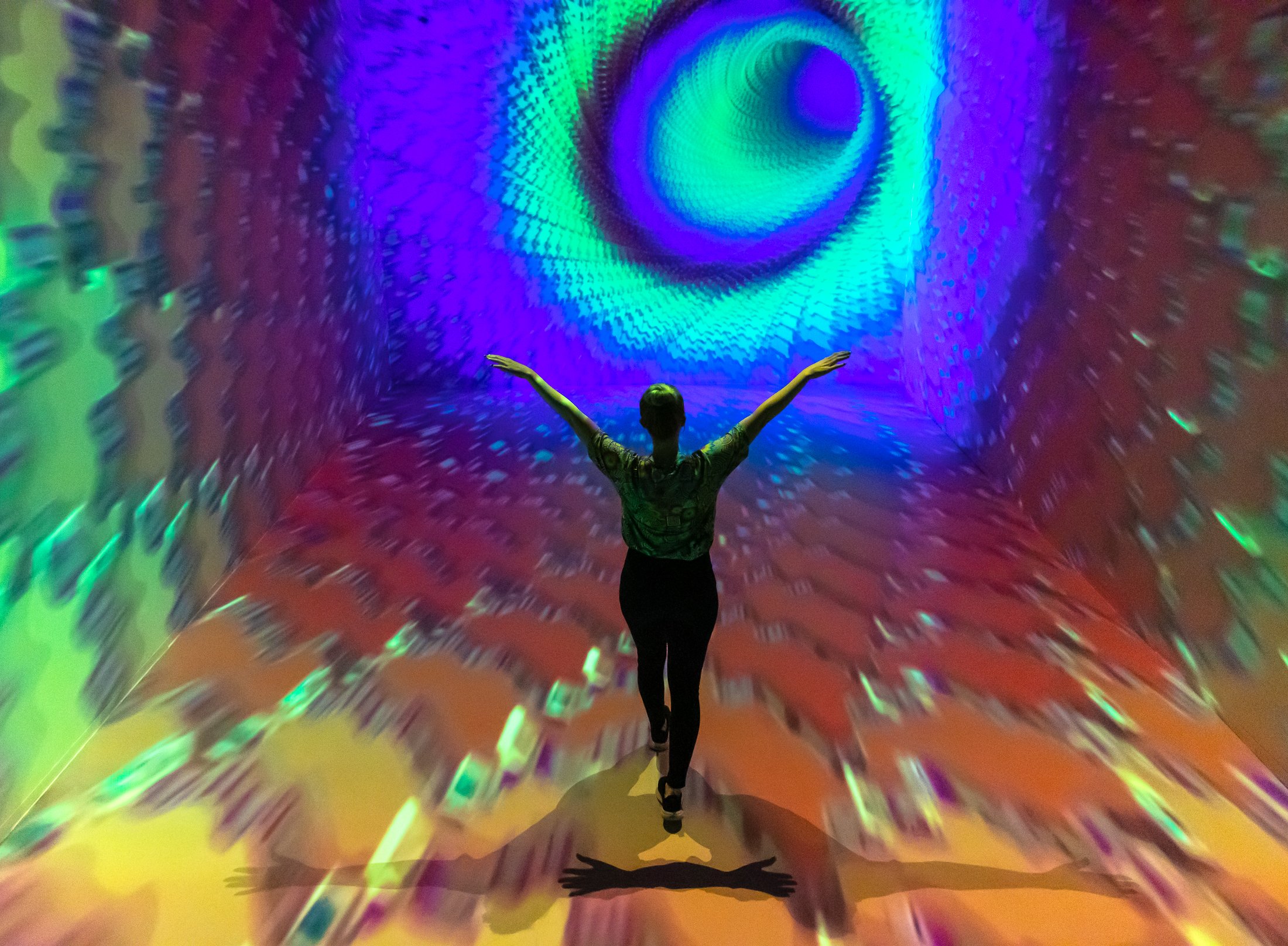 Person standing in a colorful, immersive digital art installation with arms outstretched, surrounded by swirling, vibrant, psychedelic patterns.
