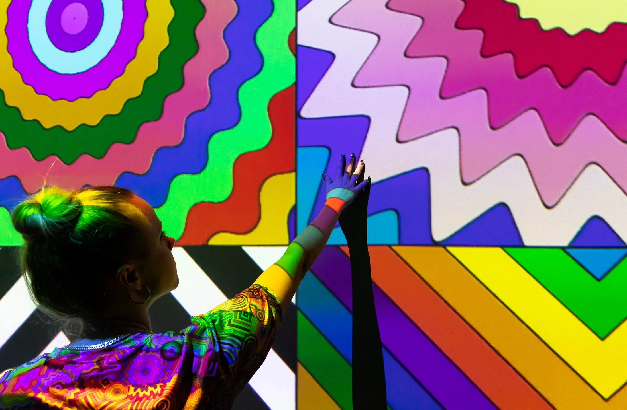 Person with rainbow-colored clothing and hair interacting with a colorful, abstract digital art piece on a screen, featuring wavy, zigzag, and striped patterns in bright colors.