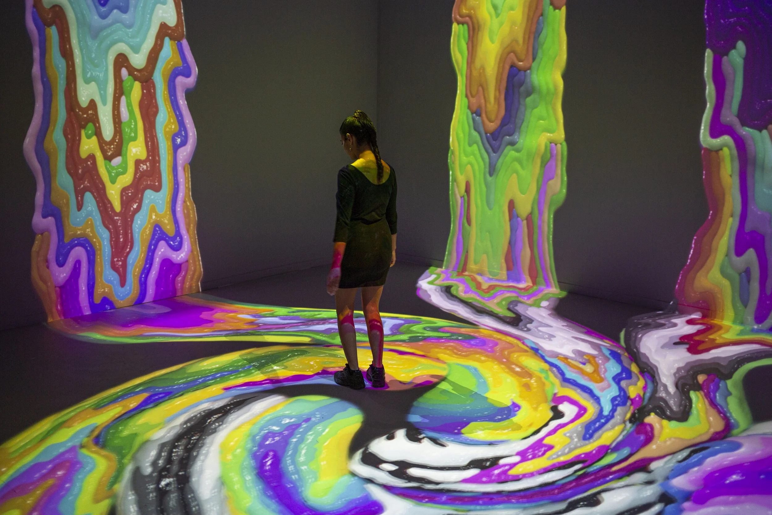 A woman standing in an immersive digital art exhibit with swirling, colorful patterns projected on the walls, floor, and her legs.