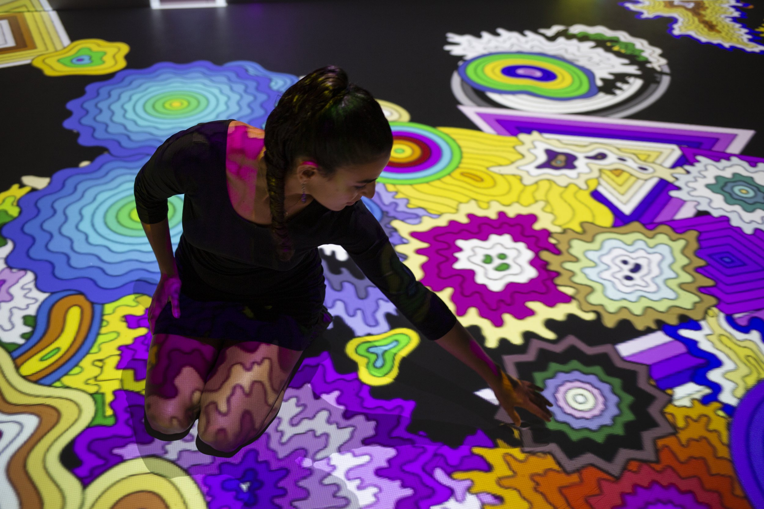 A person in a black outfit kneeling on a black floor, reaching out to touch a colorful abstract digital art projected onto the floor, featuring various vibrant geometric shapes and patterns.