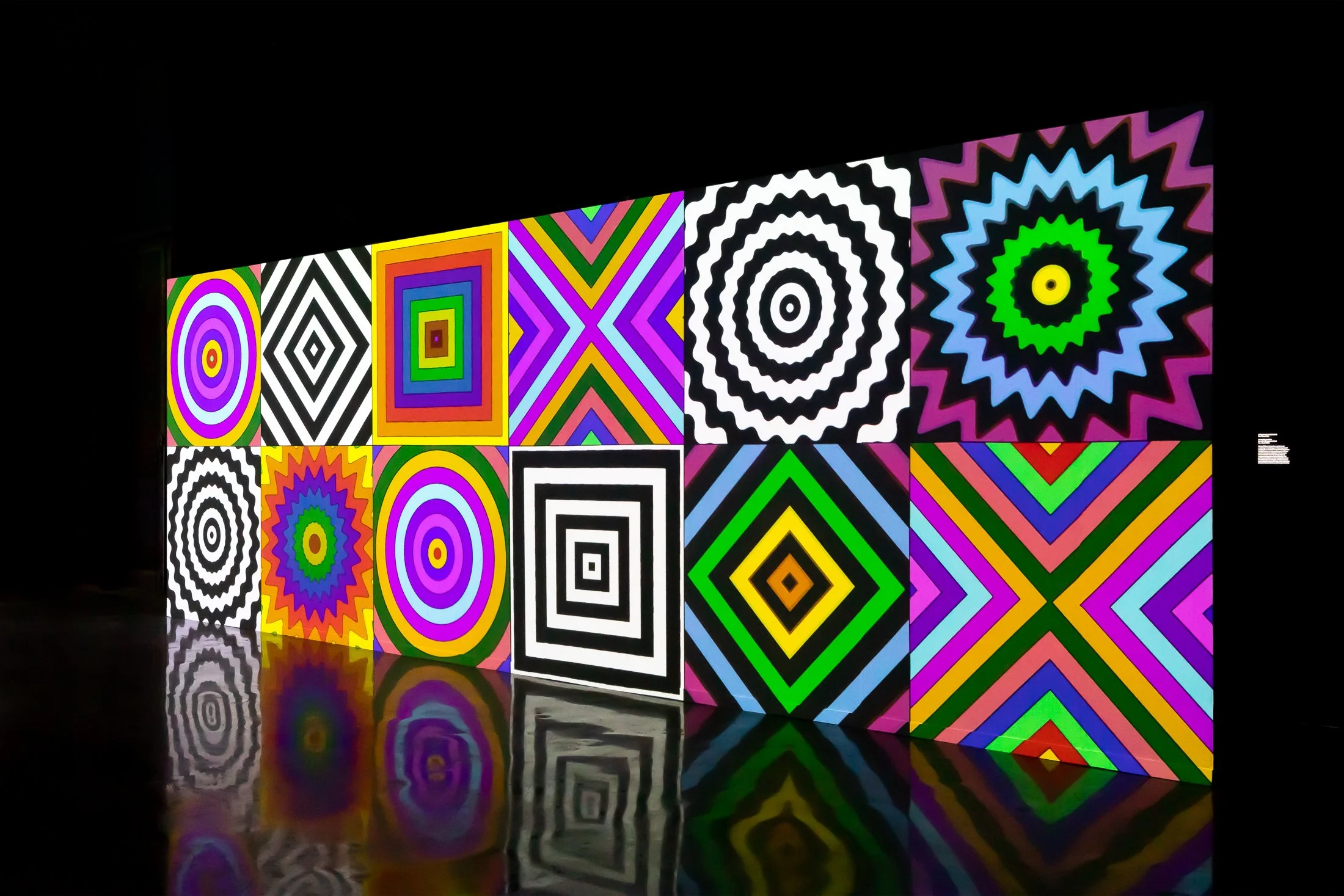 Colorful geometric patterns displayed on illuminated panels, featuring concentric circles, diamonds, and star shapes in vibrant colors such as purple, yellow, green, black, and white, reflected on the shiny surface below.