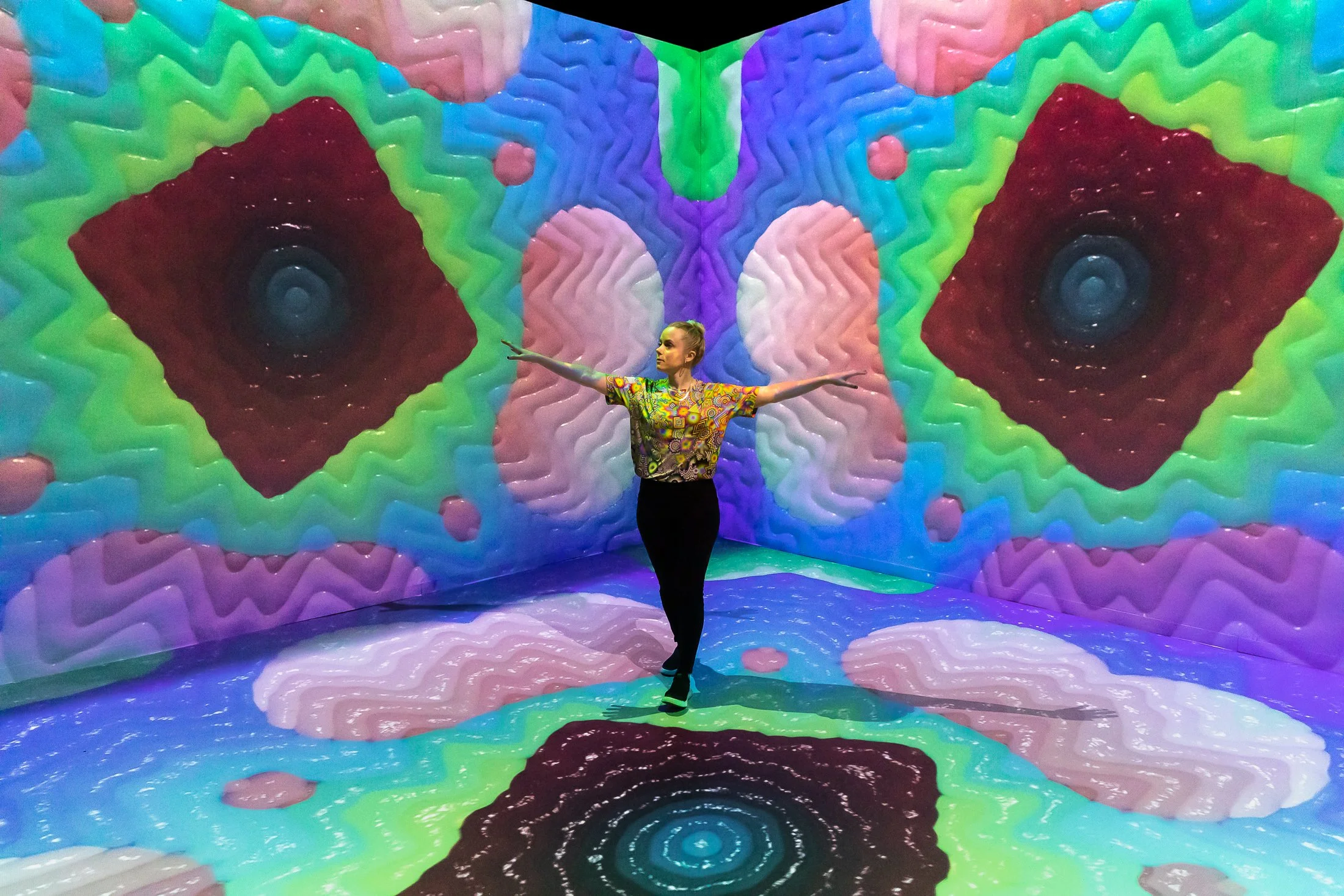 A woman posing with arms outstretched in a colorful, psychedelic room with digital art projected on the walls and floor, featuring symmetrical patterns and vibrant colors.