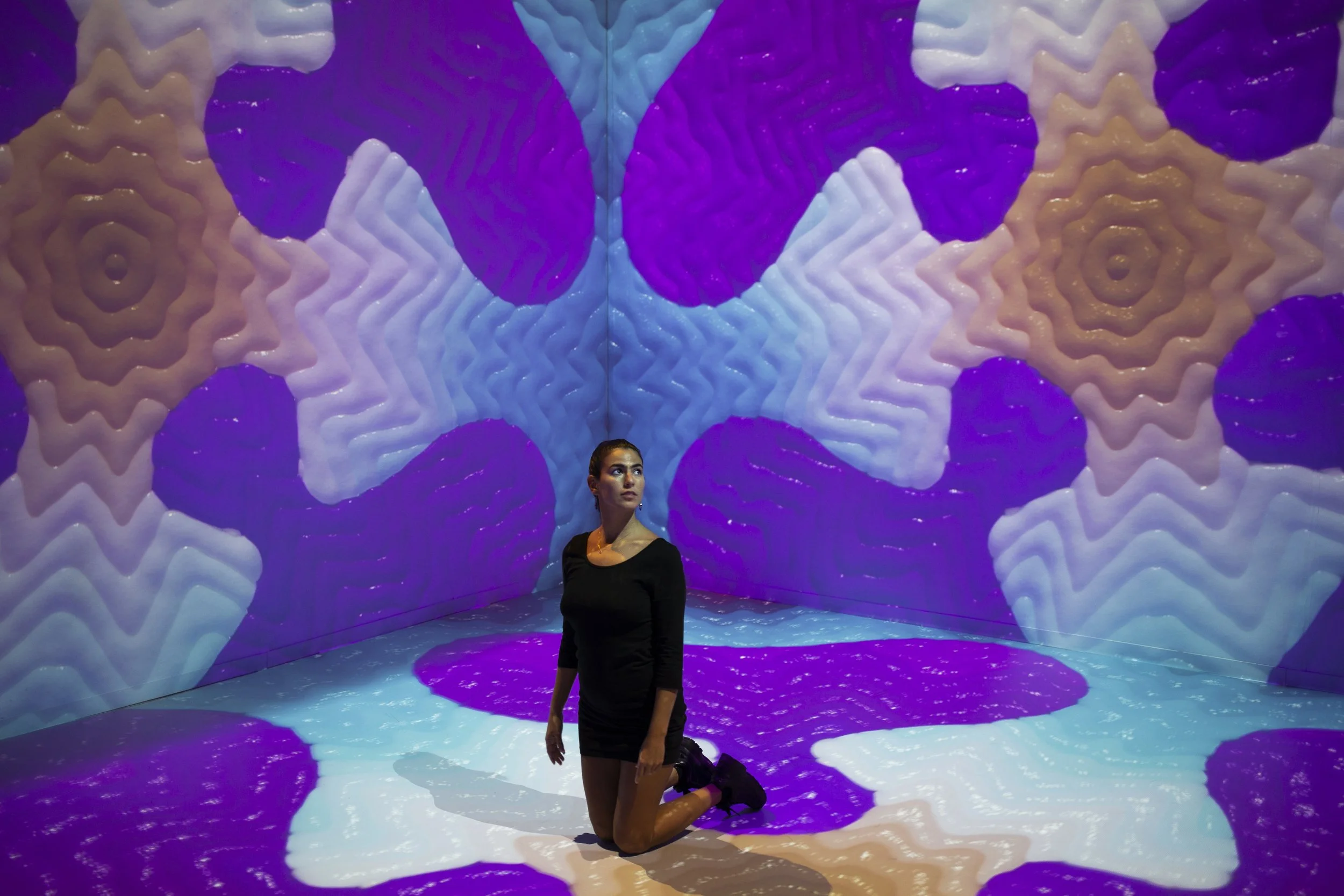 A woman is kneeling on the floor surrounded by an immersive digital art installation with colorful, patterned projections on the walls and floor.