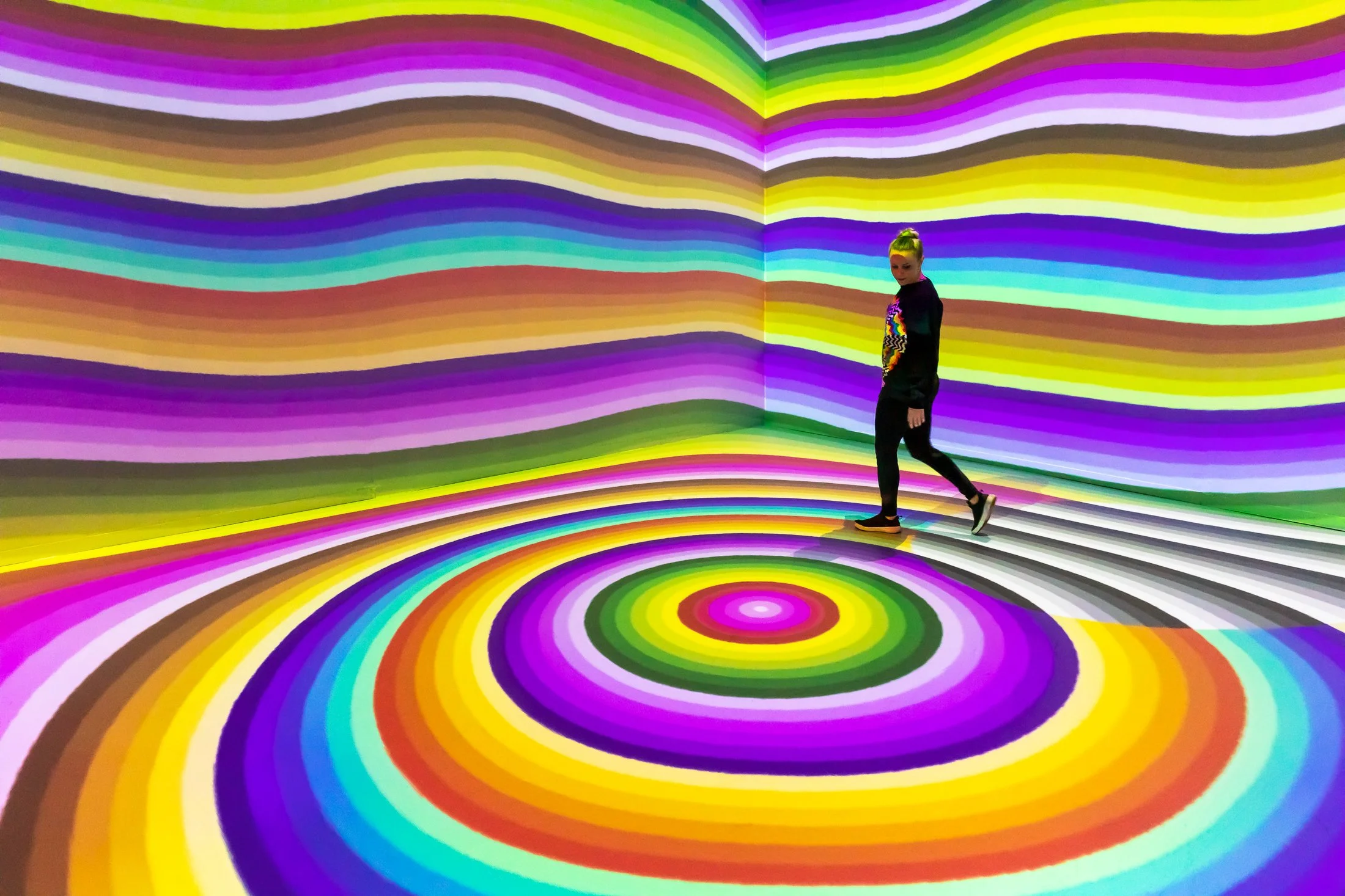 A person walking through a vibrant, colorful room with swirling rainbow patterns on the walls, floor, and ceiling.