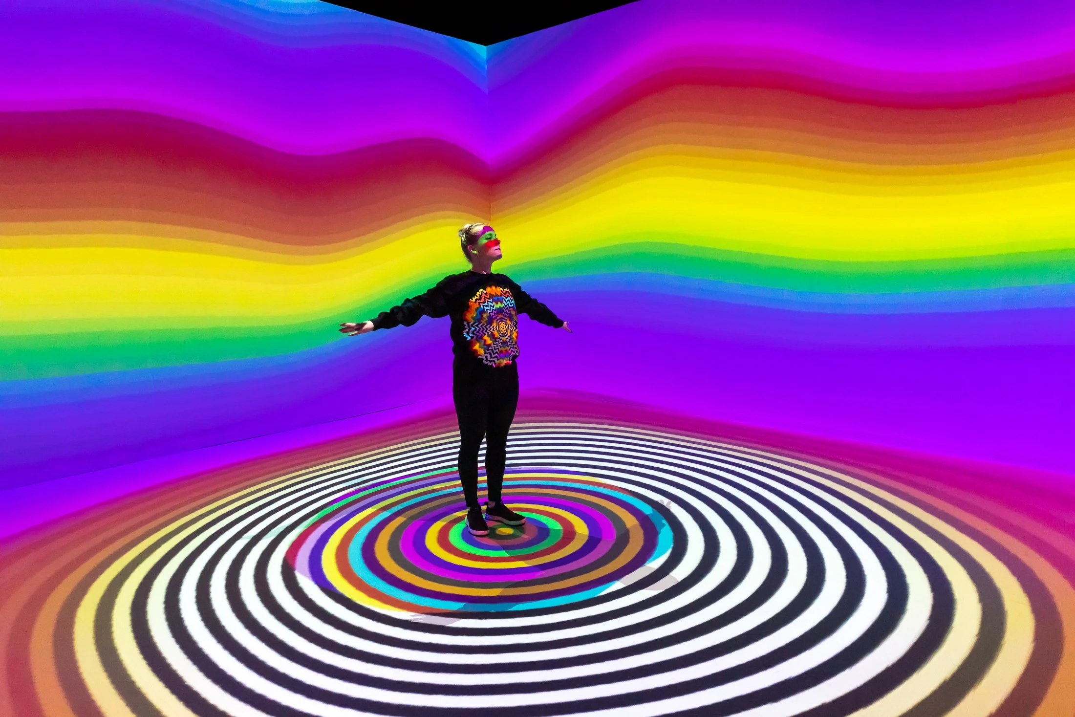 Person standing with arms outstretched in a room with vibrant, colorful concentric circles on the floor and rainbow stripes on the walls, creating an optical illusion.