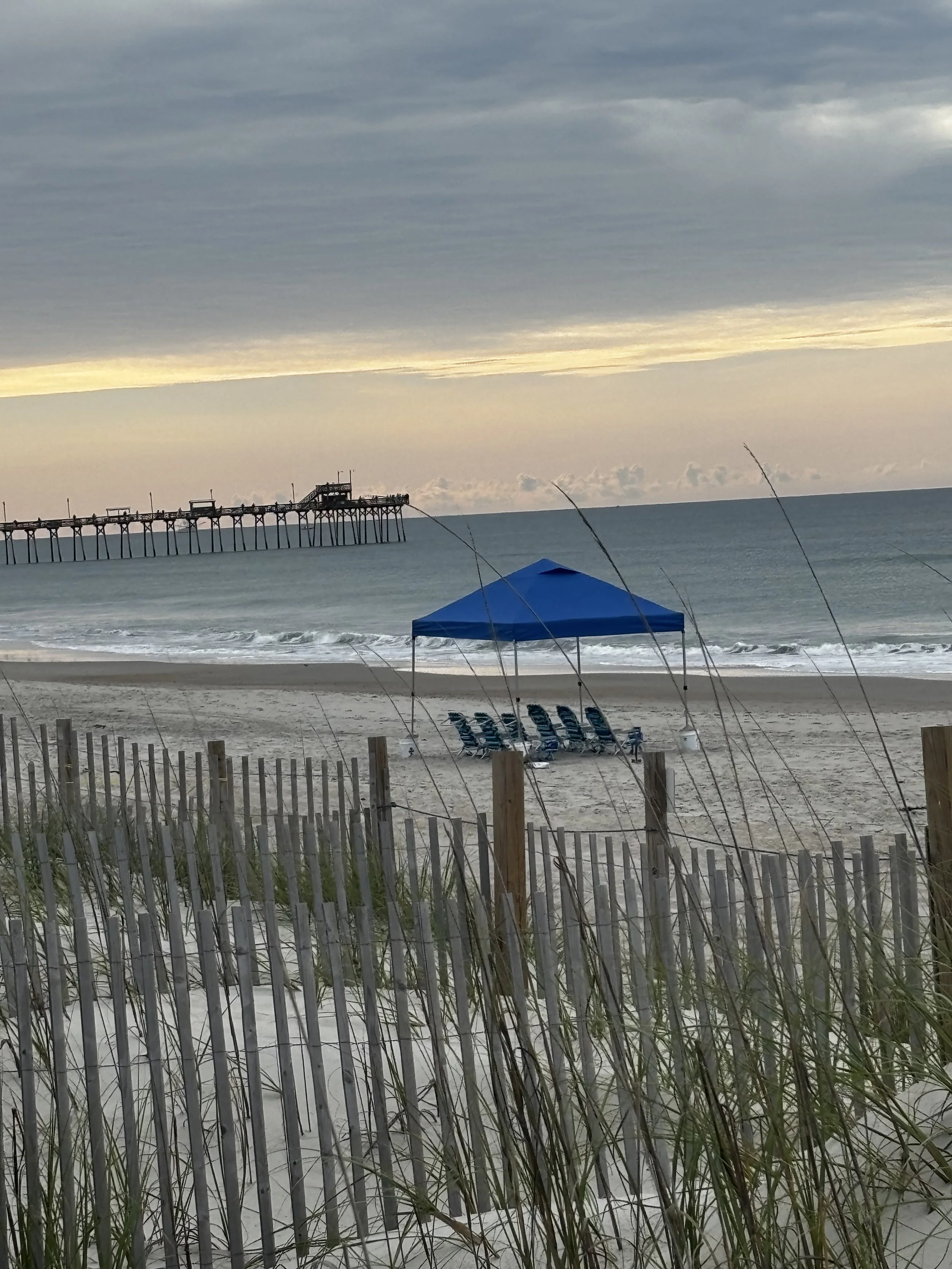 Empty beach with a blue canopy tent, several lounge chairs, and a pier in the distance during cloudy weather.