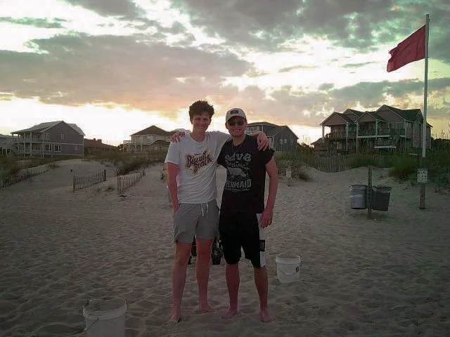 Two young men standing on a sandy beach at sunset, smiling with arms around each other's shoulders. One is wearing a white T-shirt and gray shorts, the other is wearing a black T-shirt and black shorts. Behind them, there are houses and a red flag on a pole.