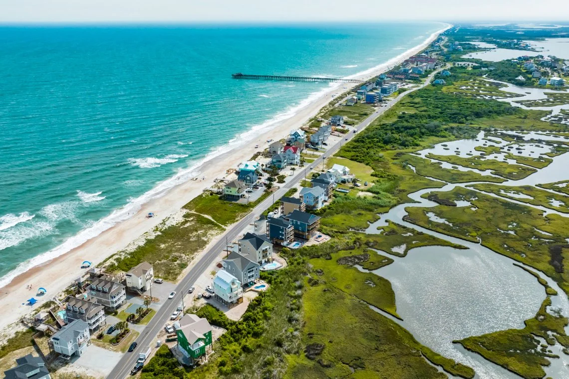 Aerial view of a coastal area with houses along the beach and marshlands on the other side.
