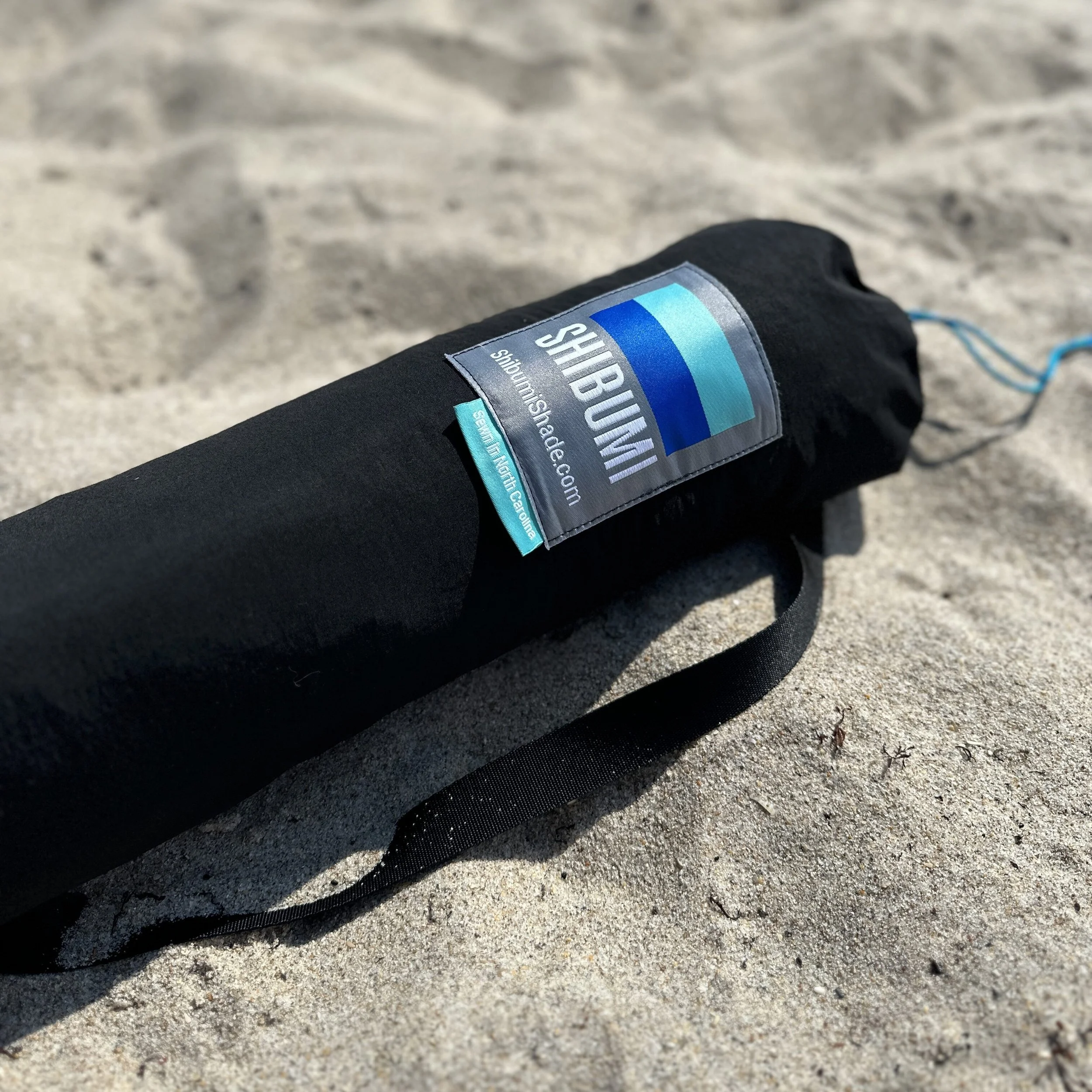 A black carrying case for a shadow tent lying on sandy ground with a label that says "SHADOW" and a website URL.