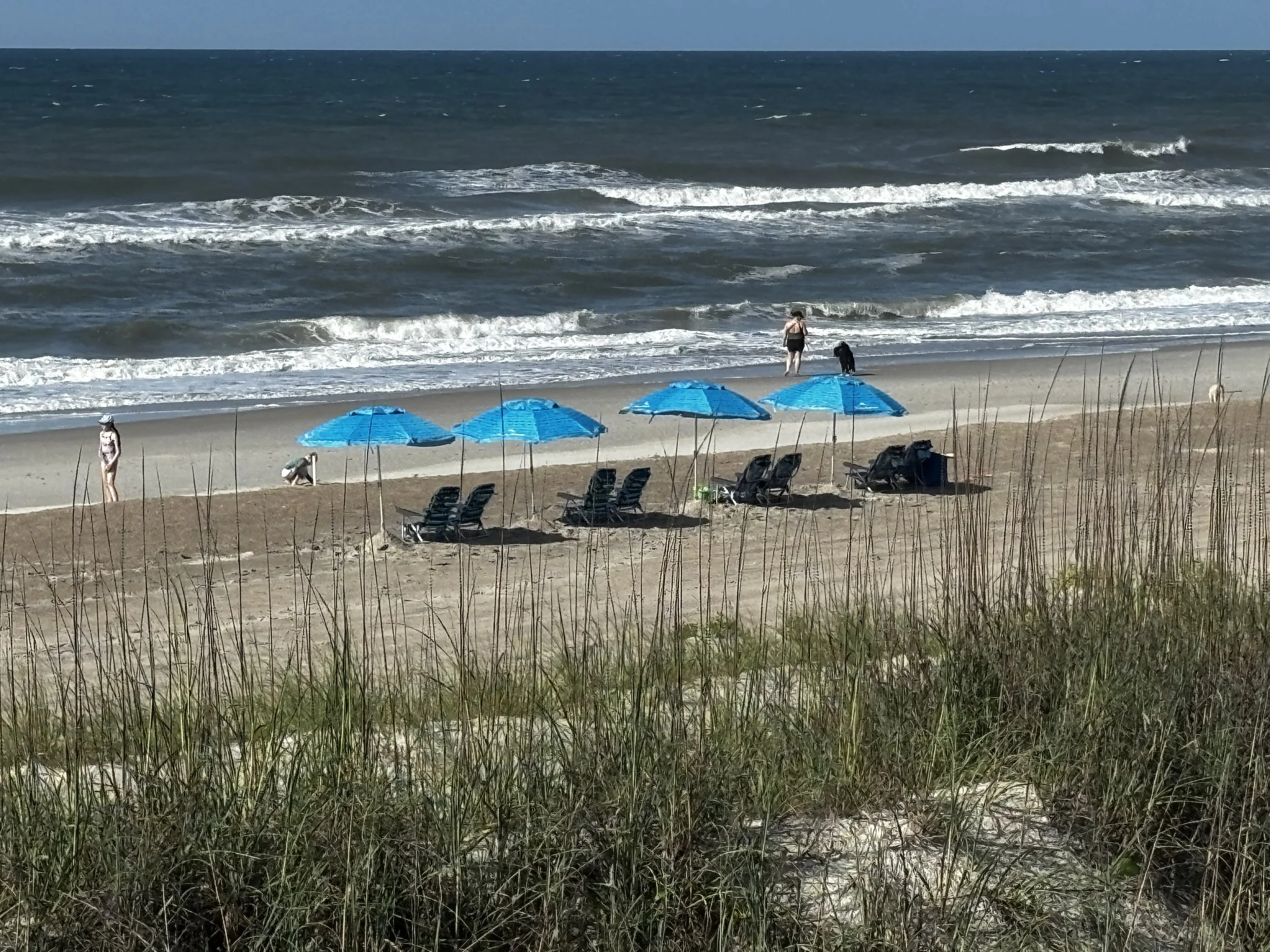 Beach with blue umbrellas, black lounge chairs, and people walking along the shore. The ocean has small waves and the sky is partly cloudy.