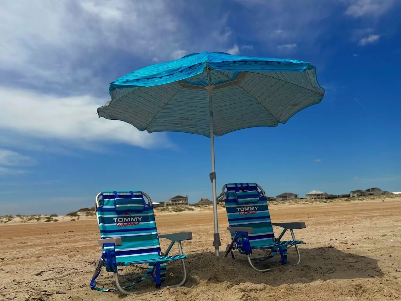 Two blue and white striped beach chairs under a large beach umbrella on sandy beach with houses in the distance.