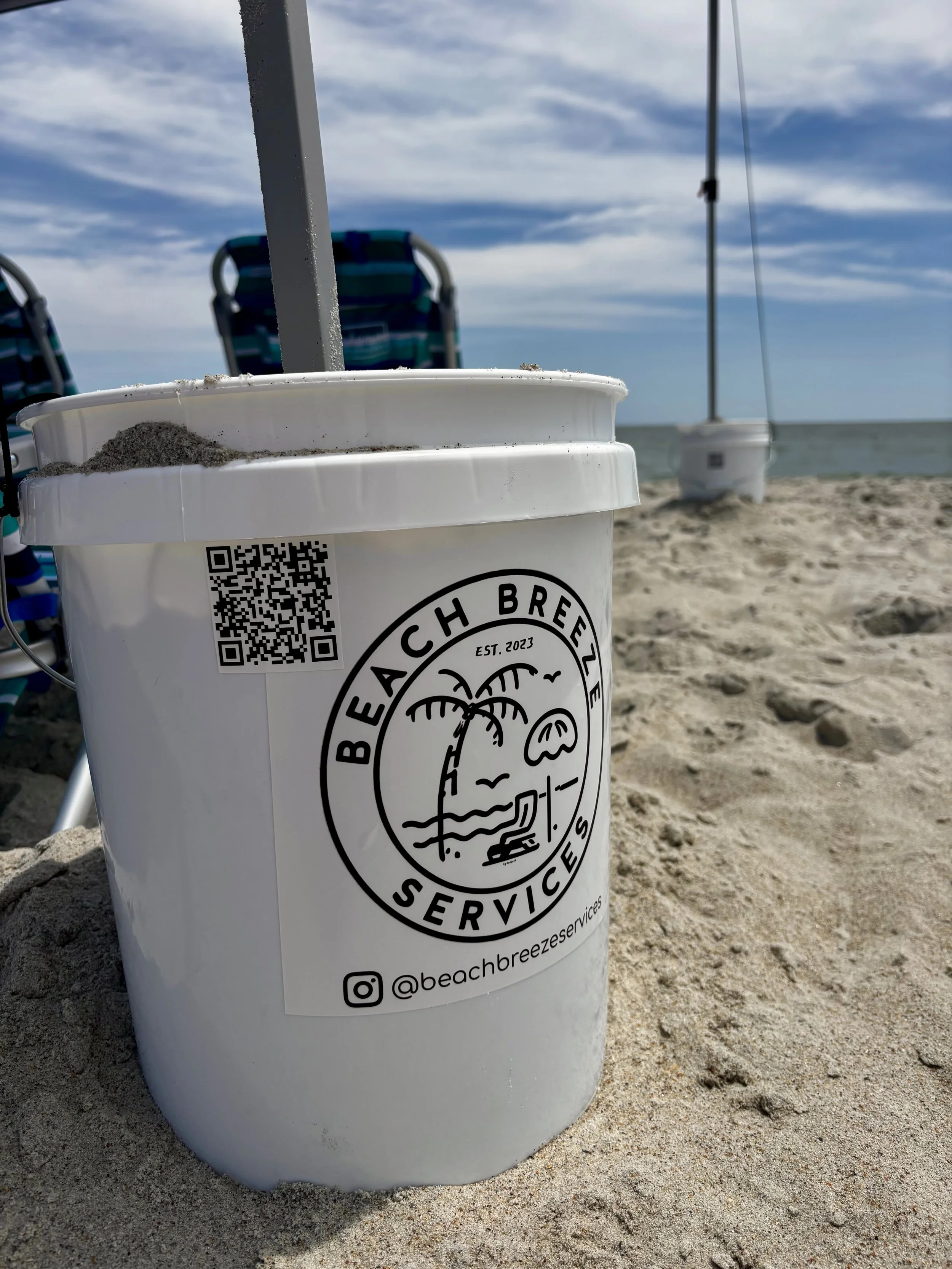 White bucket with a logo for Beach Breeze Services on a sandy beach, with an umbrella and beach chairs in the background, and the ocean and sky visible.