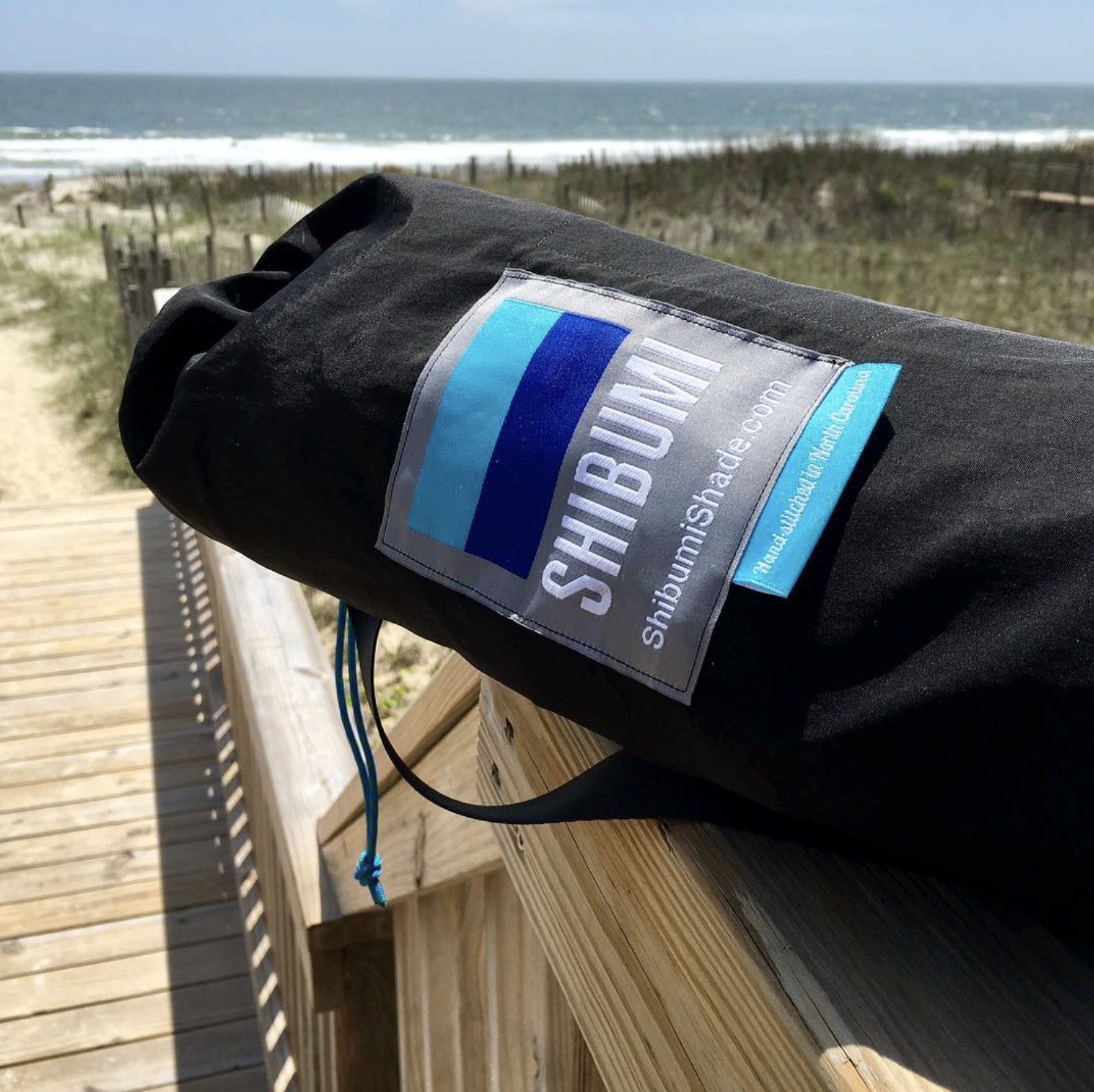 A black Shibumi pack resting on a wooden handrail on a boardwalk by the beach with sand dunes and ocean in the background.