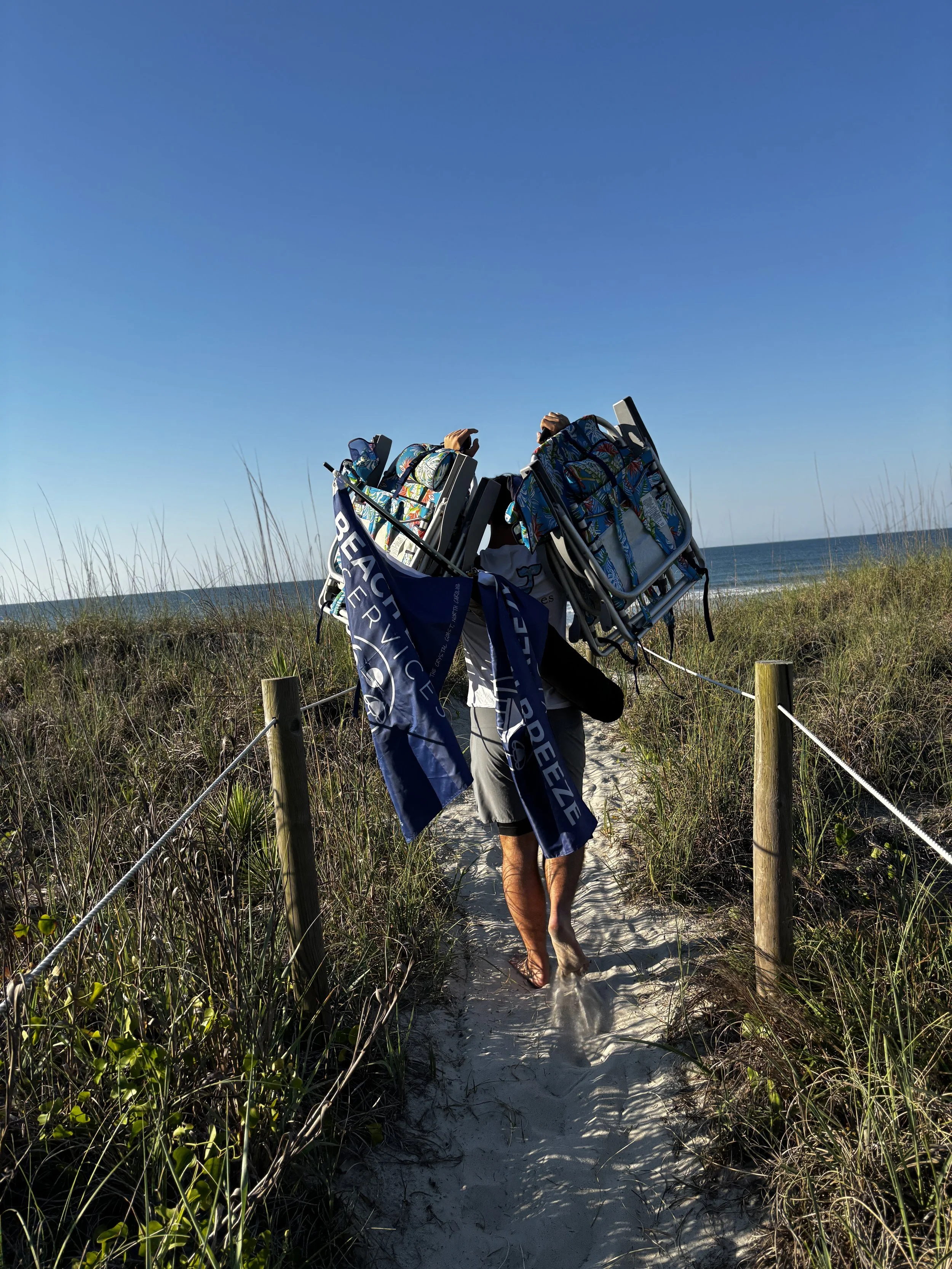 A person Walking on a beach trail carrying multiple folded chairs, near dunes and the ocean under a clear blue sky.