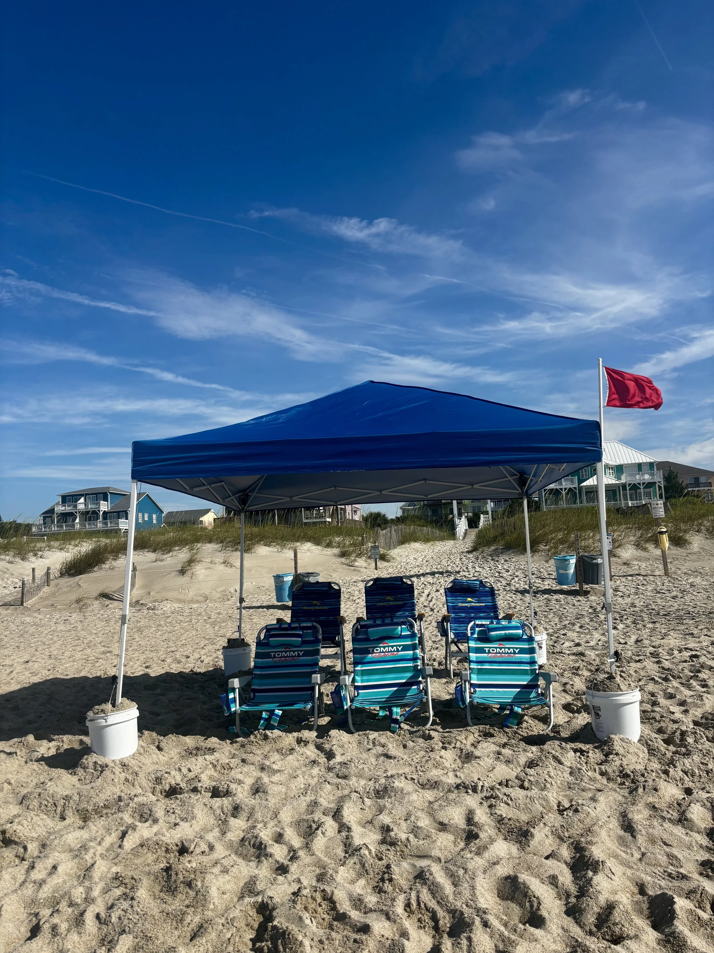 Beach scene with a blue canopy, six striped beach chairs, a red flag, and surrounding houses under a partly cloudy sky.