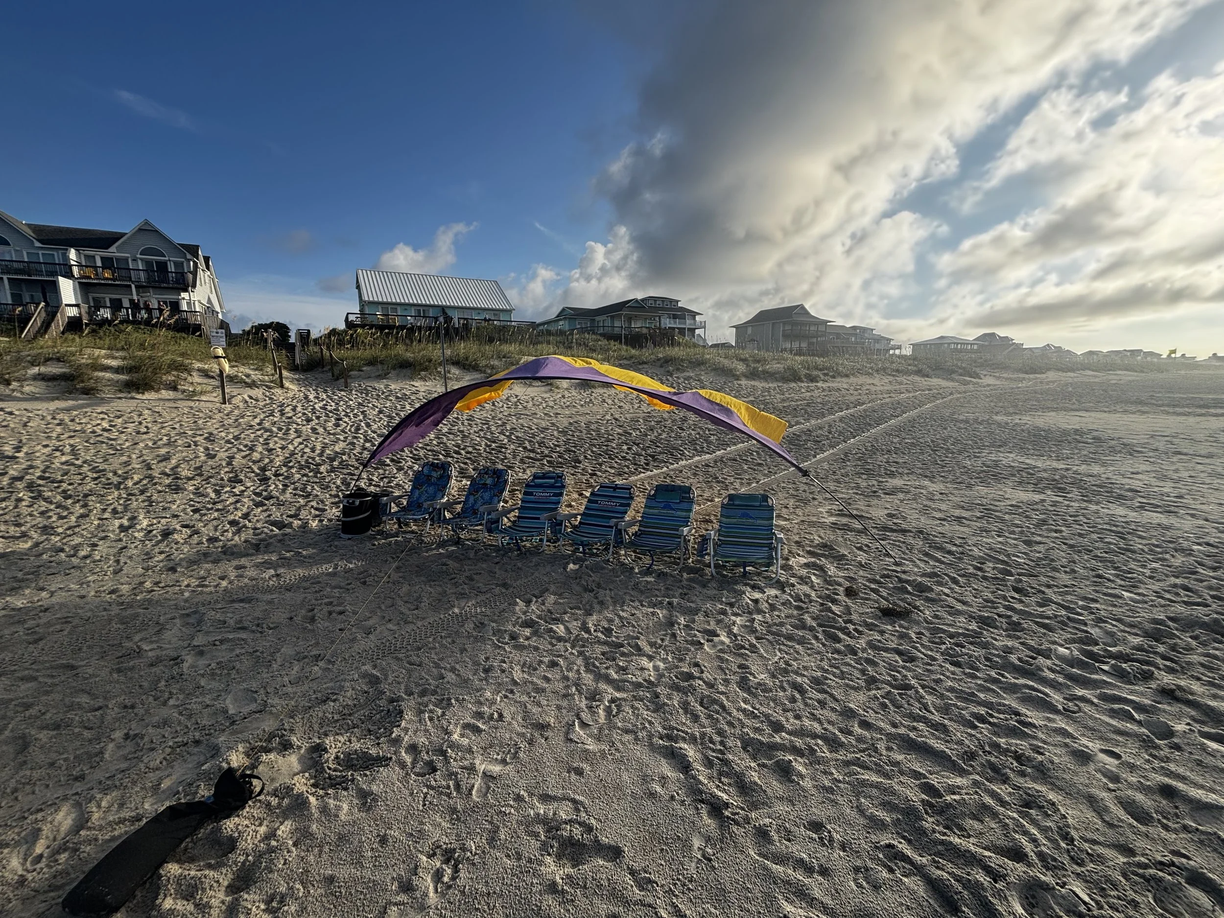 Beach scene with six lounge chairs under a purple and yellow shade sail, with houses on sandy dunes in the background and a partly cloudy sky.