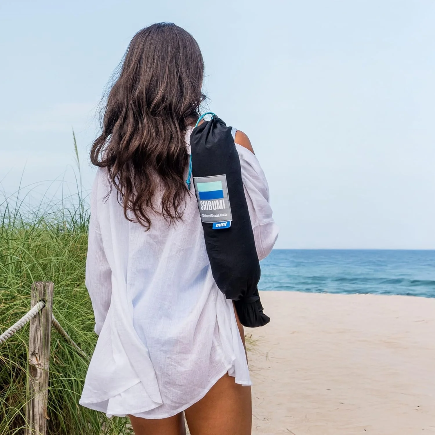 A woman standing on a beach with tall grass and a sandy path, looking at the ocean.