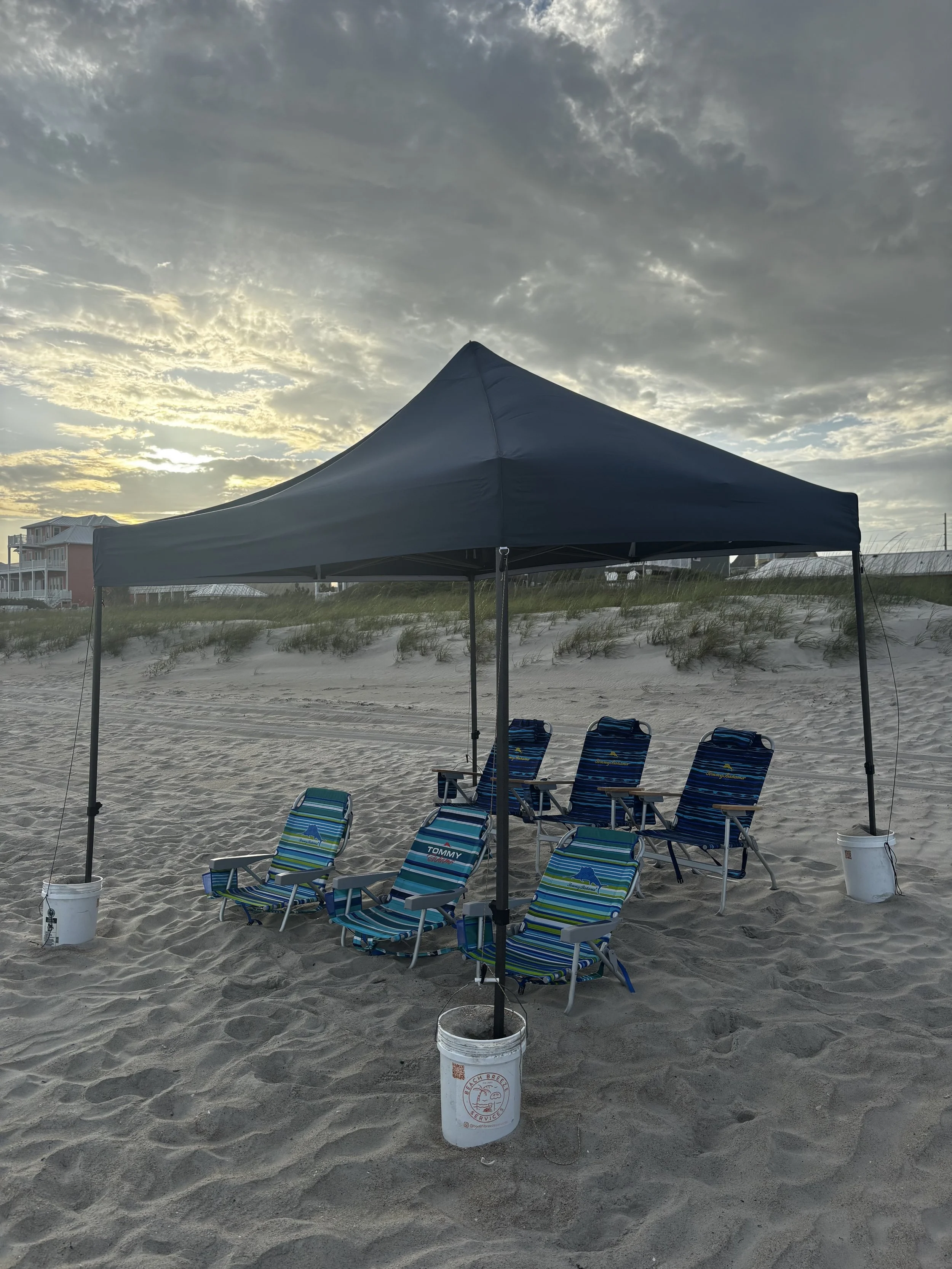 Beach tent set up on sandy beach with four chairs and two small loungers, with houses and grassy dunes in the background, under a cloudy sky.