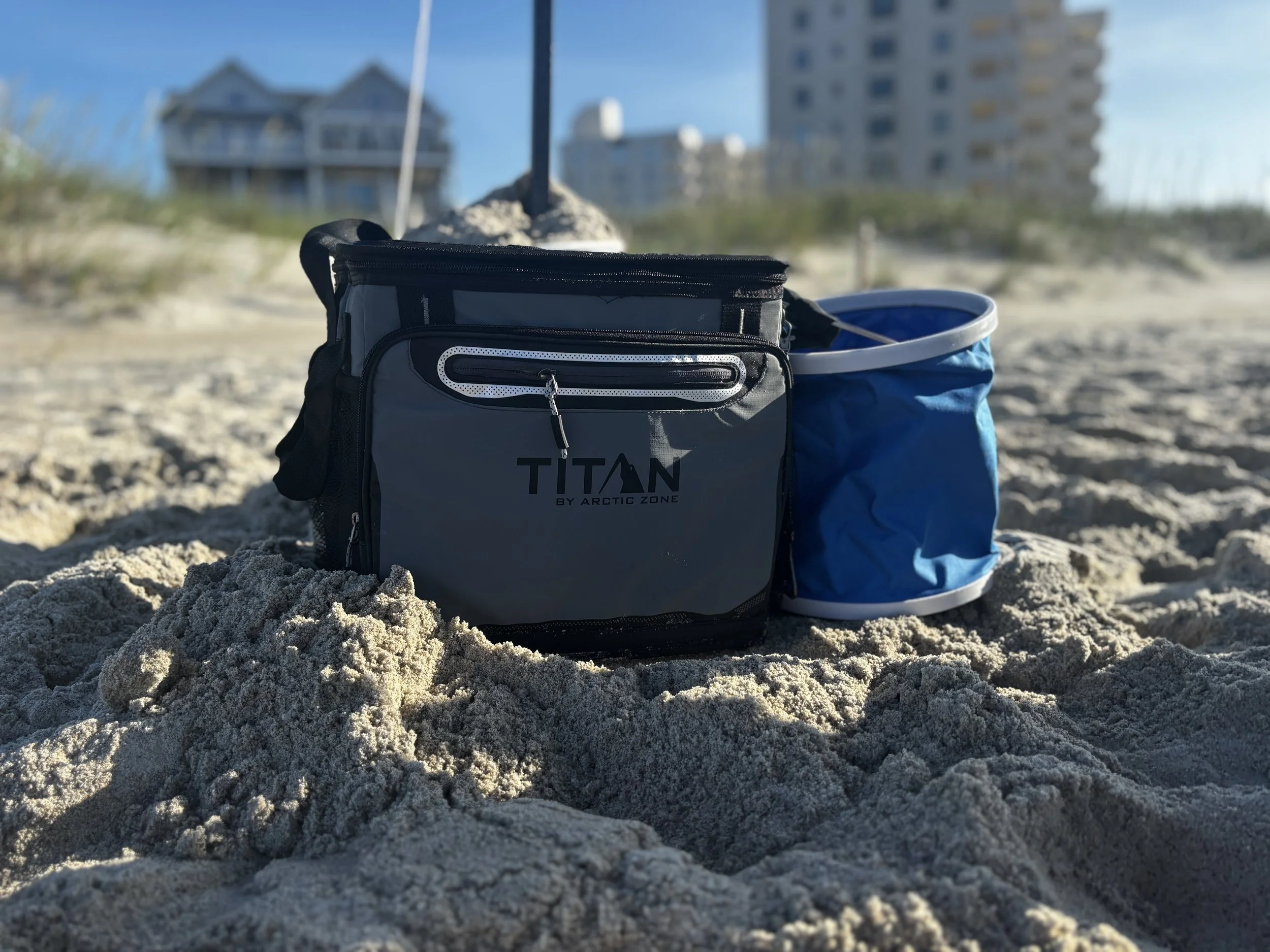 A gray and black Arctic Zone Titan cooler bag and a blue collapsible bucket set on sand at the beach with buildings and dunes in the background.
