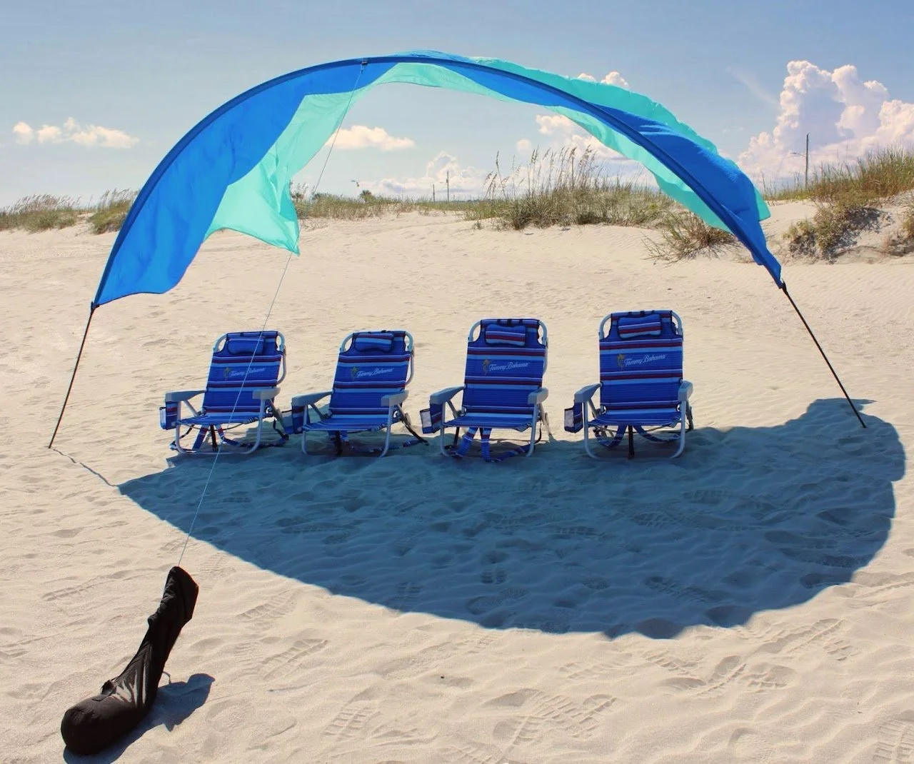 Four blue beach chairs under a blue and green umbrella on sandy beach with grass and cloudy sky in background.