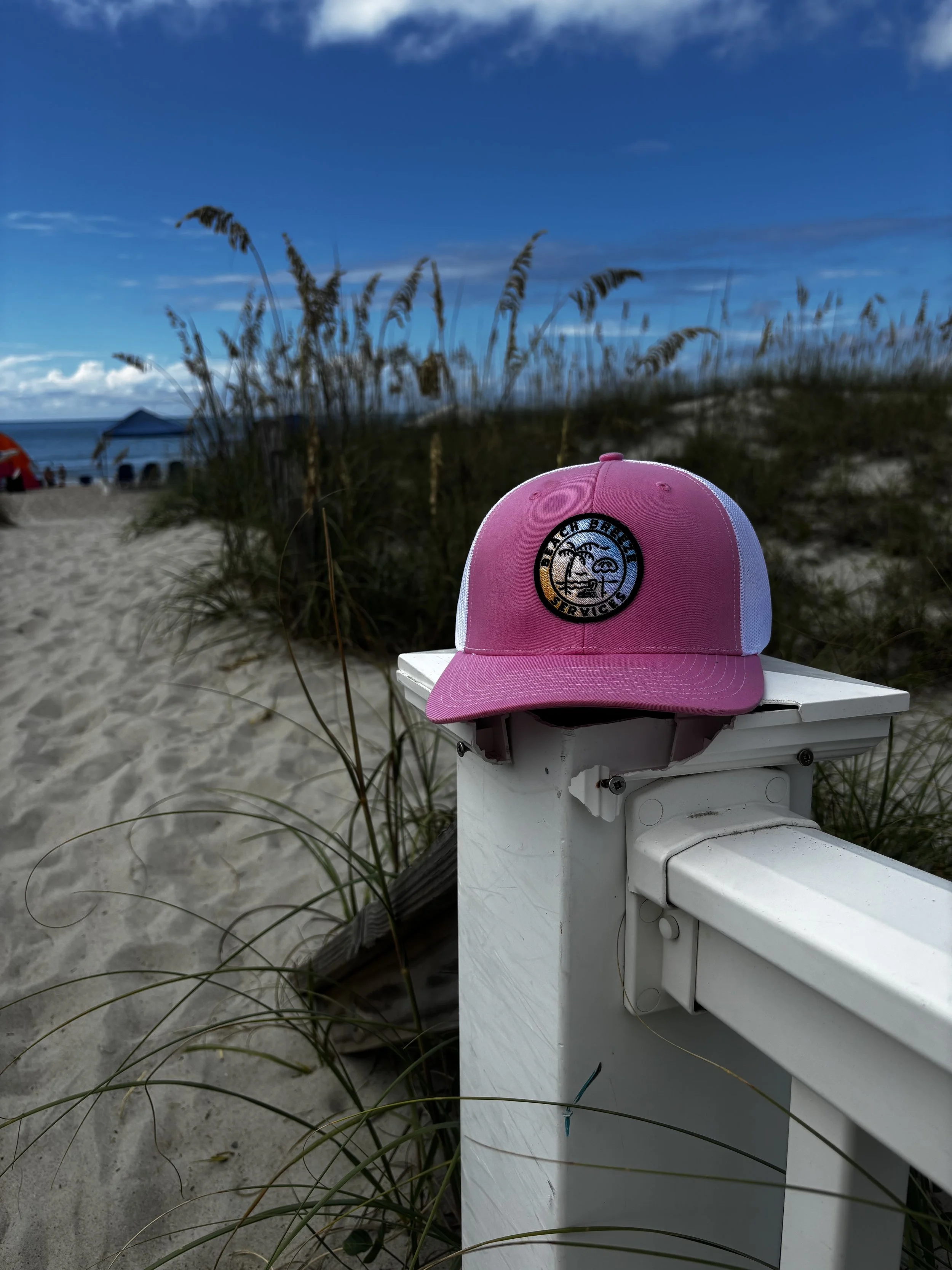 Pink and white baseball cap resting on a white fence on a sandy beach with beach grass, blue sky, and ocean in the background.