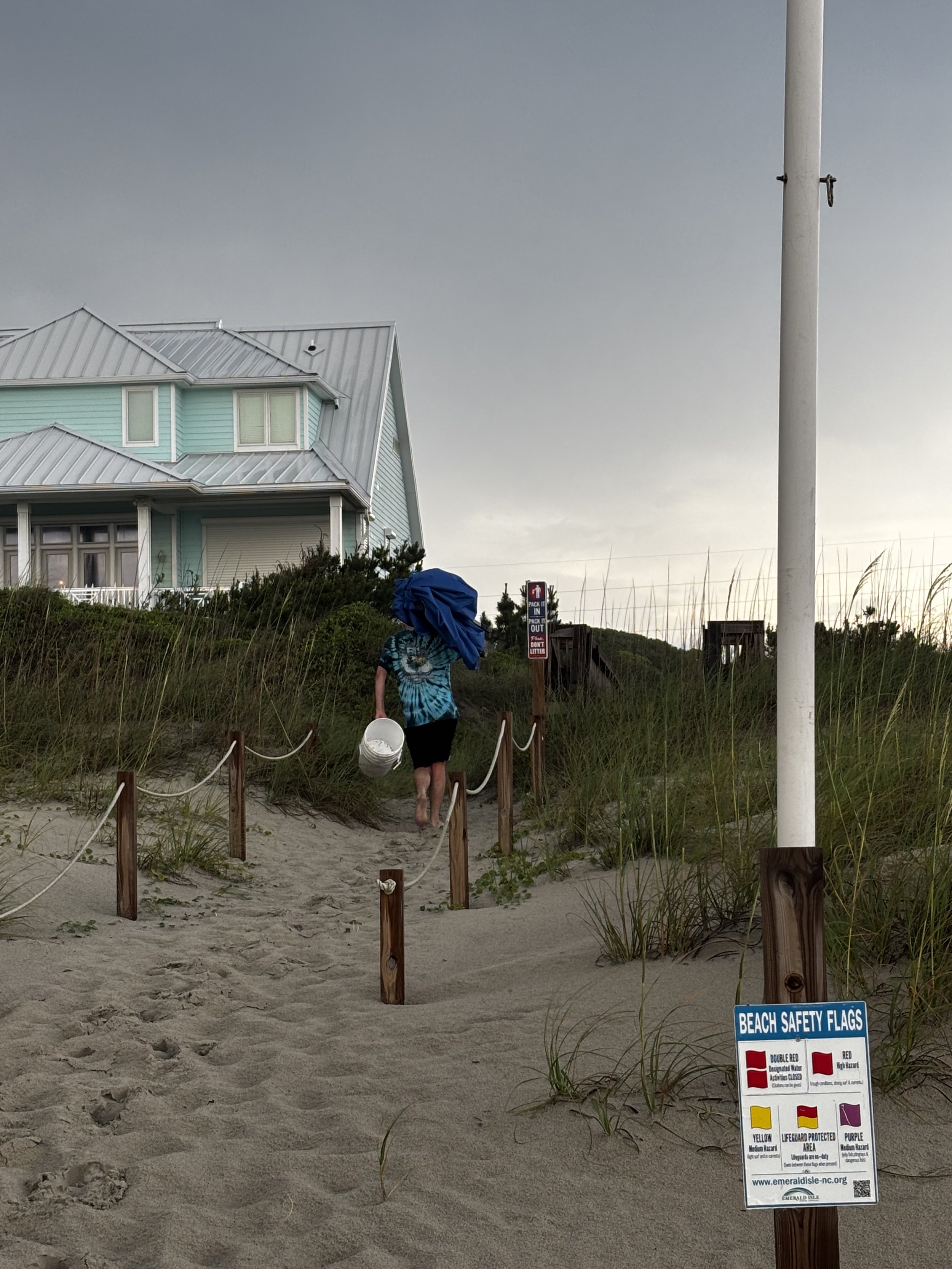 A person walking on a sandy beach path carrying a white bucket, with a large house in the background, and a sign indicating beach safety flags on the right.