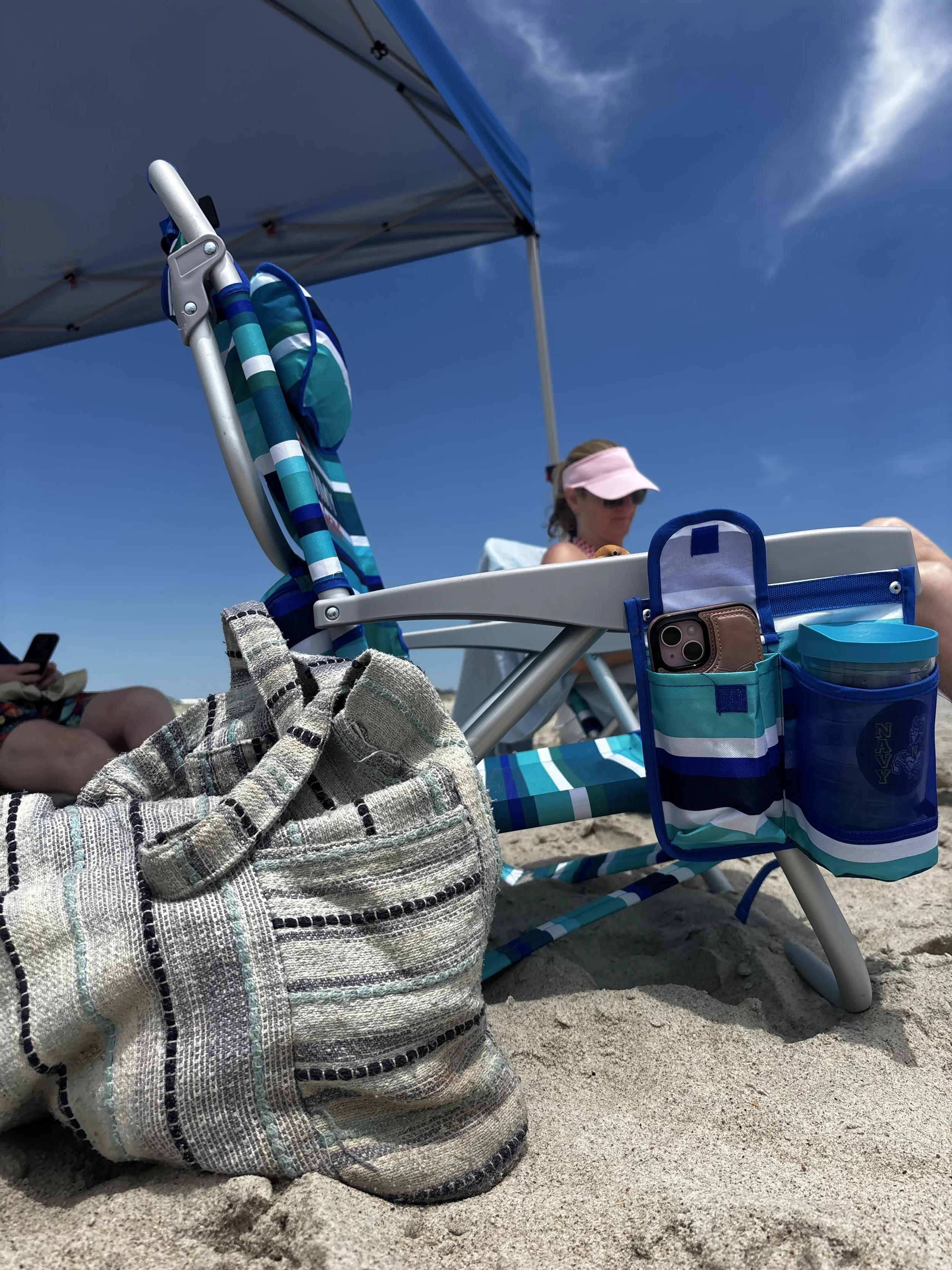 Beach scene with a striped bag on sand, a blue and white striped beach chair, a woman in a pink visor sitting under a blue canopy, and blue sky with some clouds.