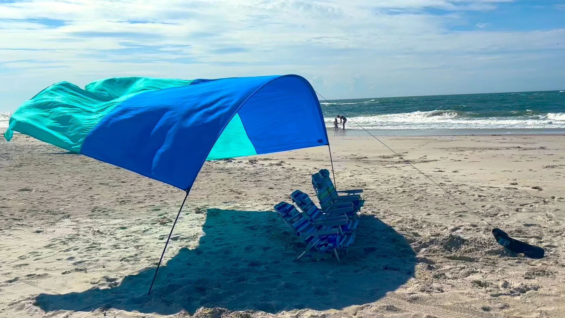 Beach scene with a blue and turquoise sunshade, multiple striped lounge chairs, and pair of people near the water in the background, with waves and a cloudy sky.