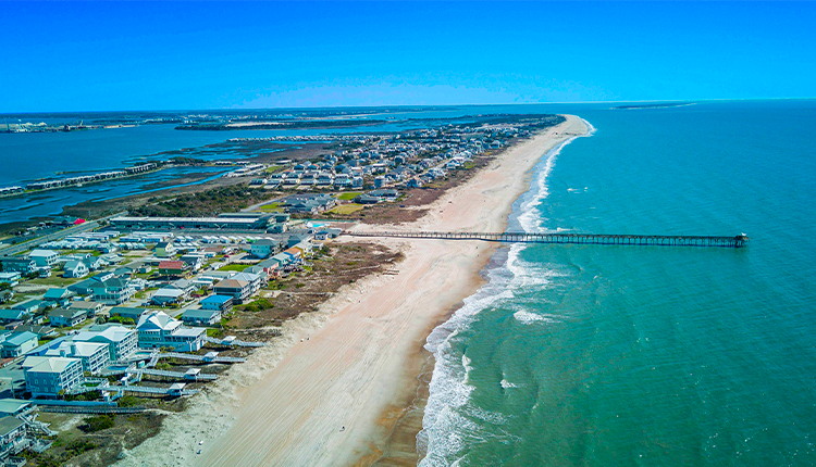 Aerial view of a beach with houses and a pier extending into the ocean on a sunny day.