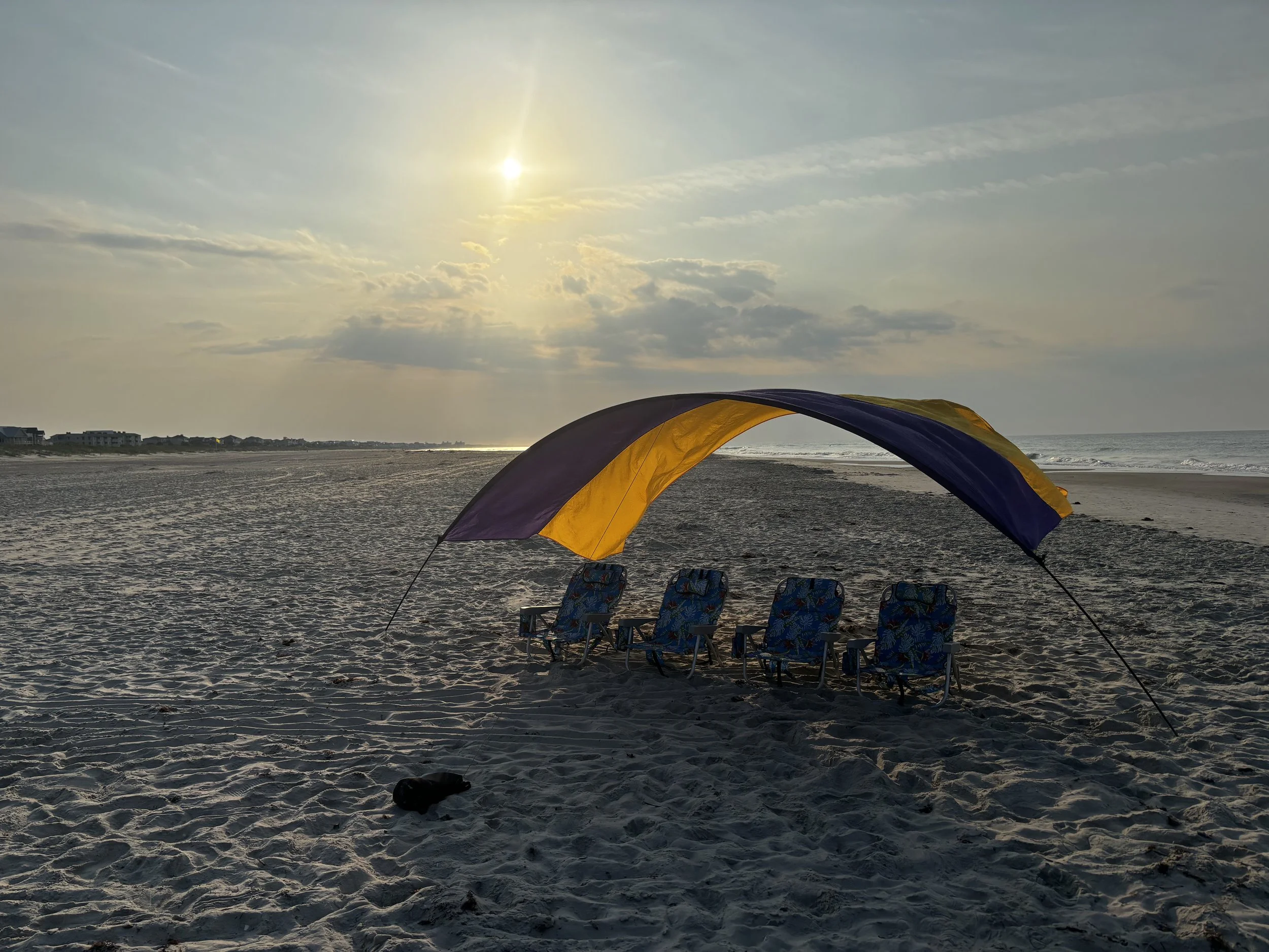 Empty beach with four chairs under a canopy, sunrise or sunset, with clouds and sun in the sky.