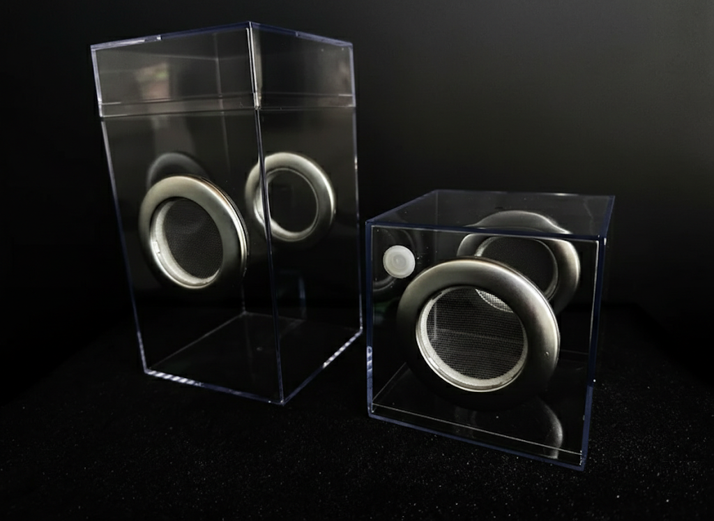Two clear acrylic jumping spider enclosures with silver mesh ventilation grommets on opposite sides, displayed against a solid black background.