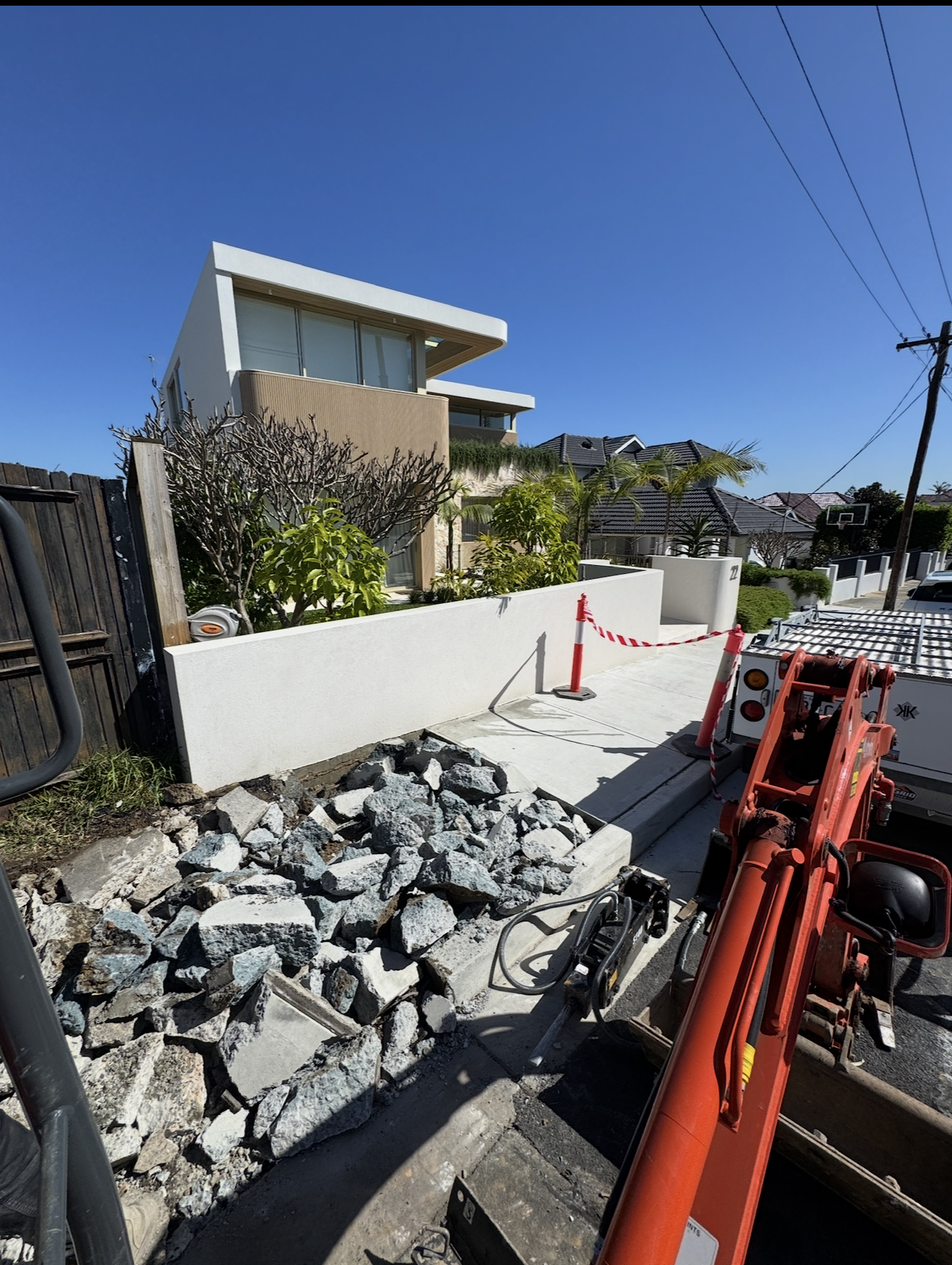 Construction site on a residential street with rocks and a partially demolished sidewalk, orange construction equipment, and houses with modern architecture and palm trees.